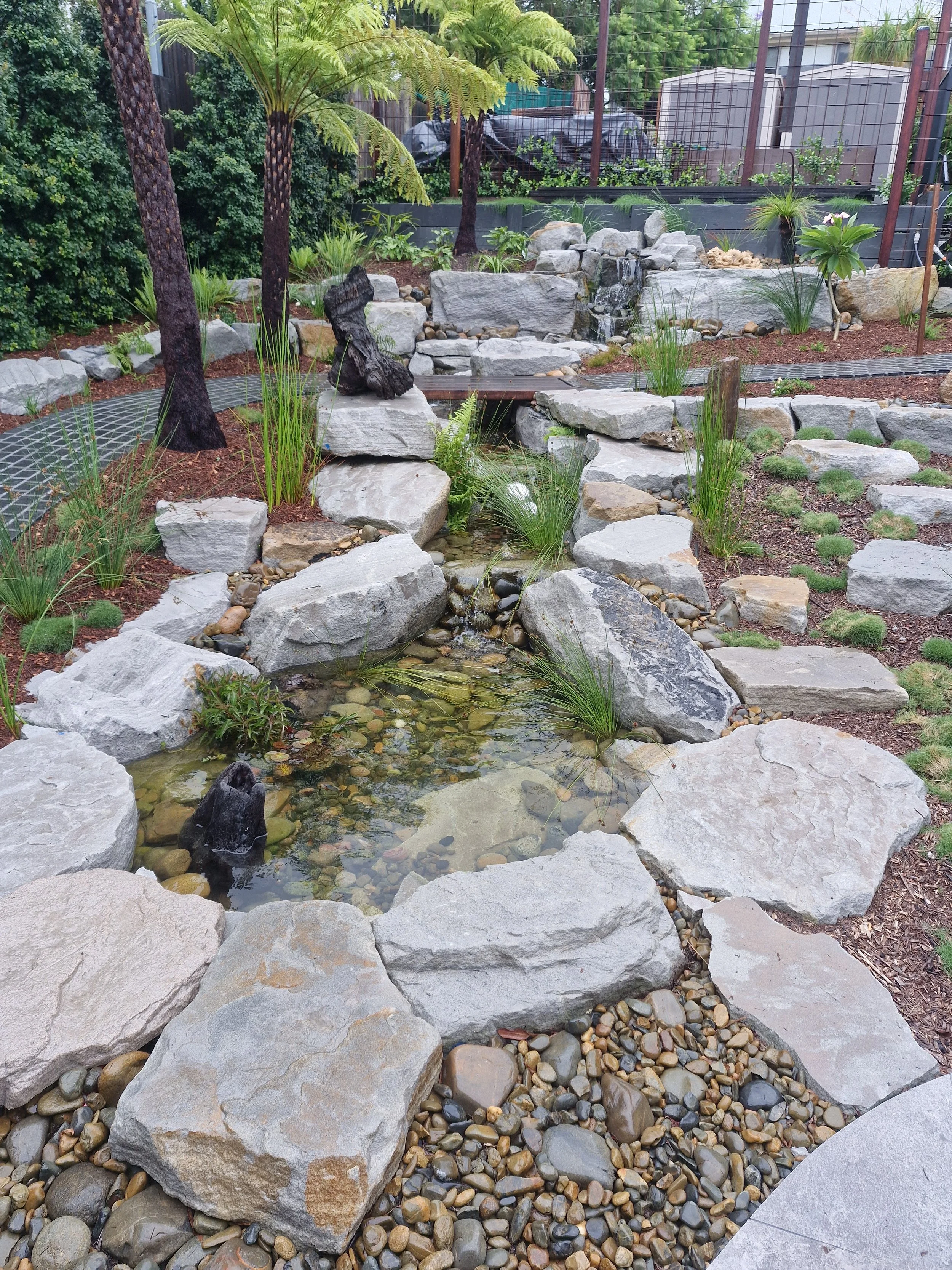 A landscaped garden with large rocks and smaller pebbles, a small flowing water feature, and lush greenery including tall trees and various plants. A wooden bridge crosses over part of the water feature, and there is a fence and some structures in the background.