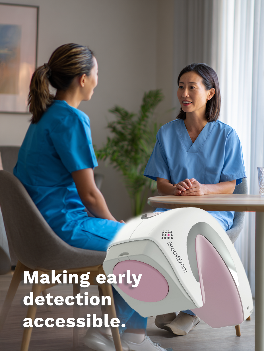 A female healthcare provider and a female patient discussing health across a round table. The white and pink hand held iBreast examination tool is overlayed on the image.