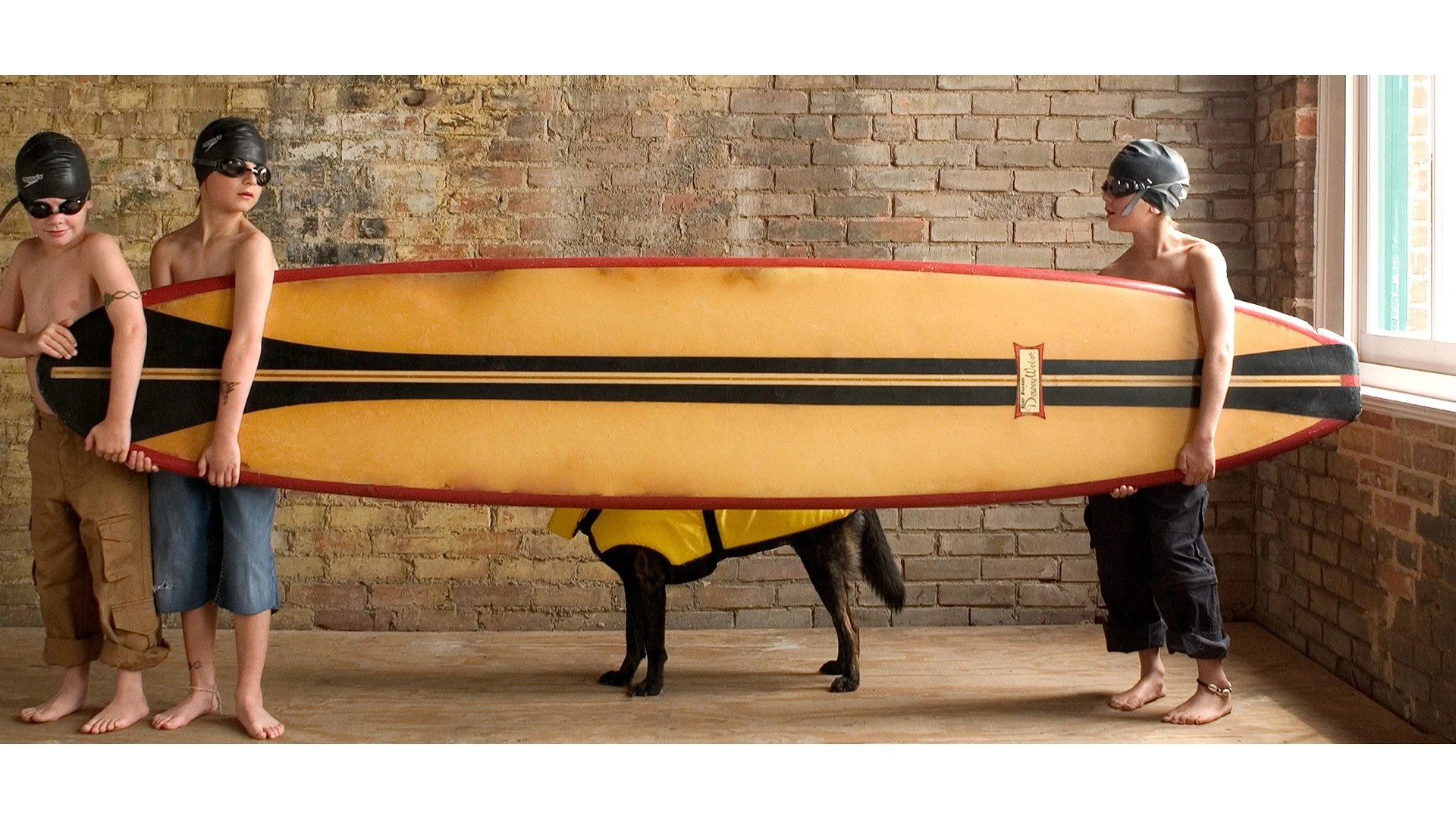 Three children with helmets and goggles, holding a large yellow surfboard with black and red accents, standing in front of a brick wall with a window to the right.