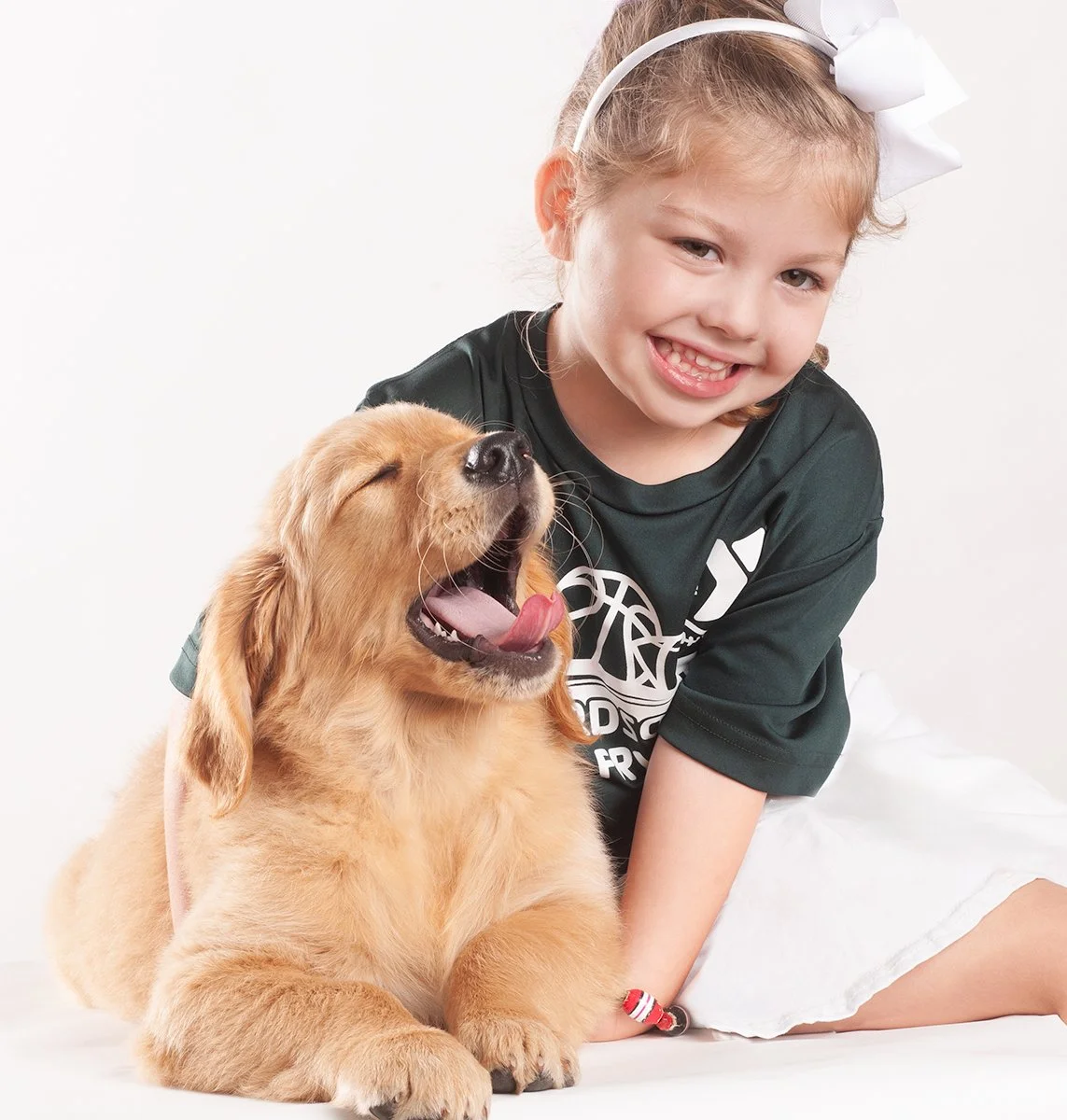 A young girl smiling and cuddling with a yawning golden retriever puppy, both sitting on a white background.