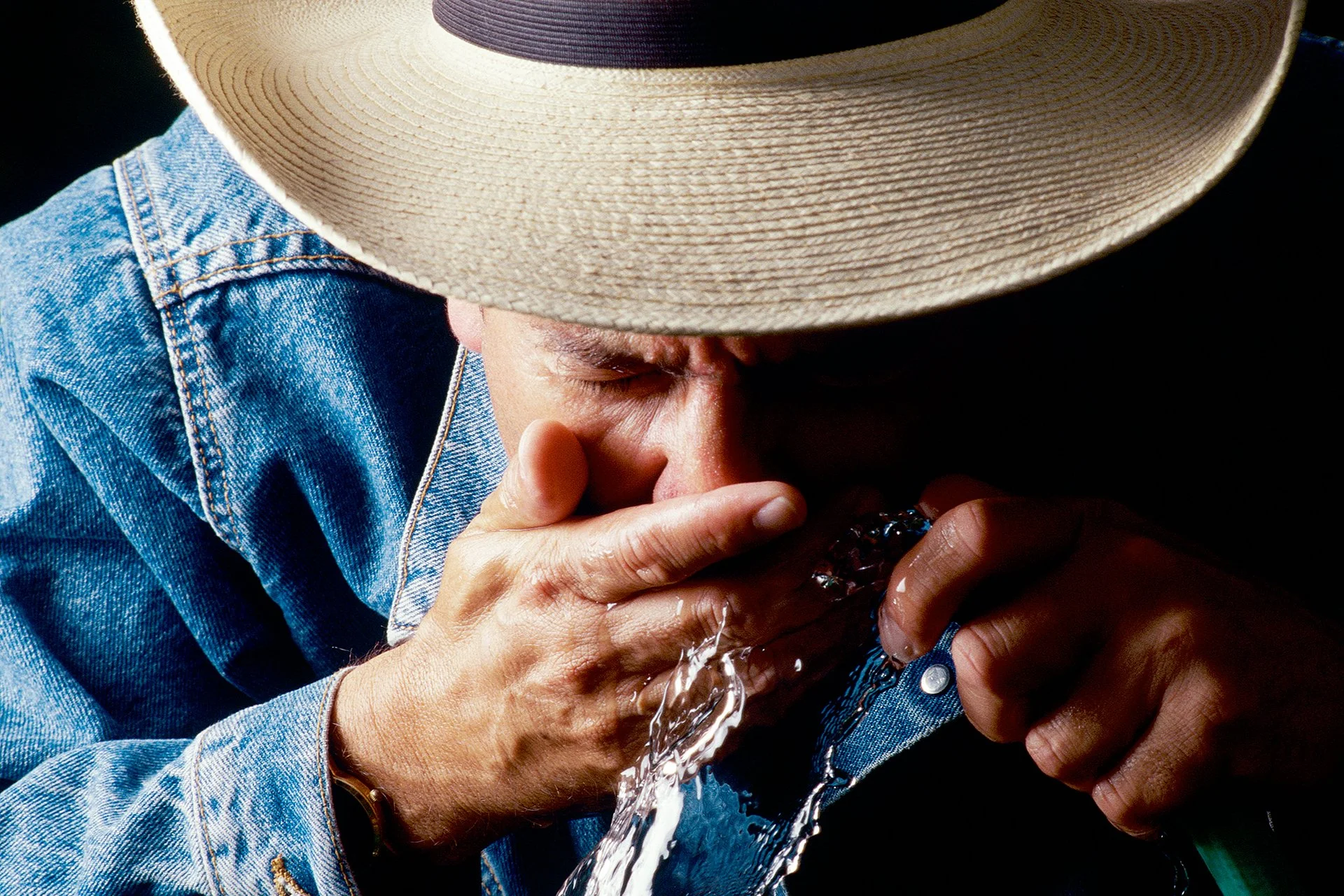 Person wearing a wide-brimmed hat and denim shirt drinking water from a bottle.