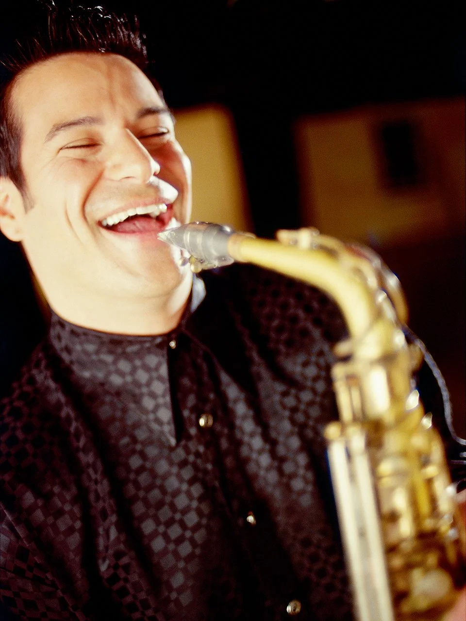 A man playing a saxophone, smiling with eyes closed, wearing a dark patterned shirt nearby a microphone.