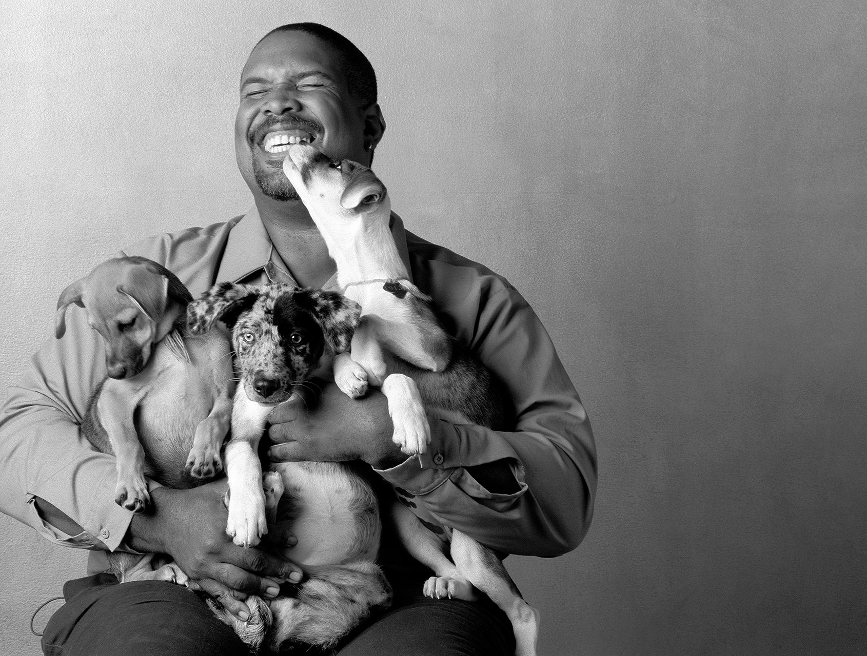 A smiling man with short hair and a goatee holding three puppies, one licking his face, against a plain background.