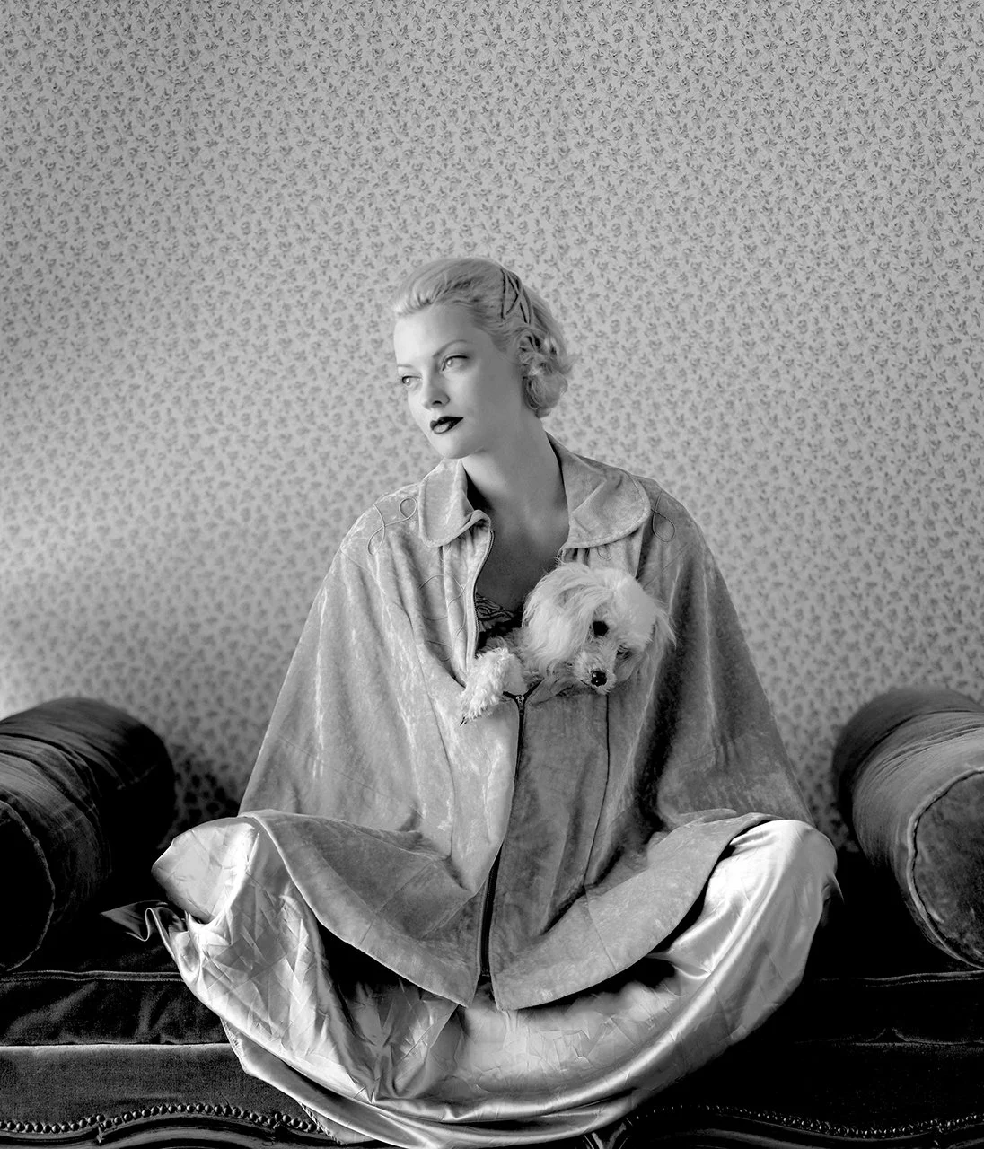 A woman sitting cross-legged on a vintage sofa holding a small plush dog. She has short, curled hair and dark lipstick, wearing a velvety jacket. The background features patterned wallpaper.