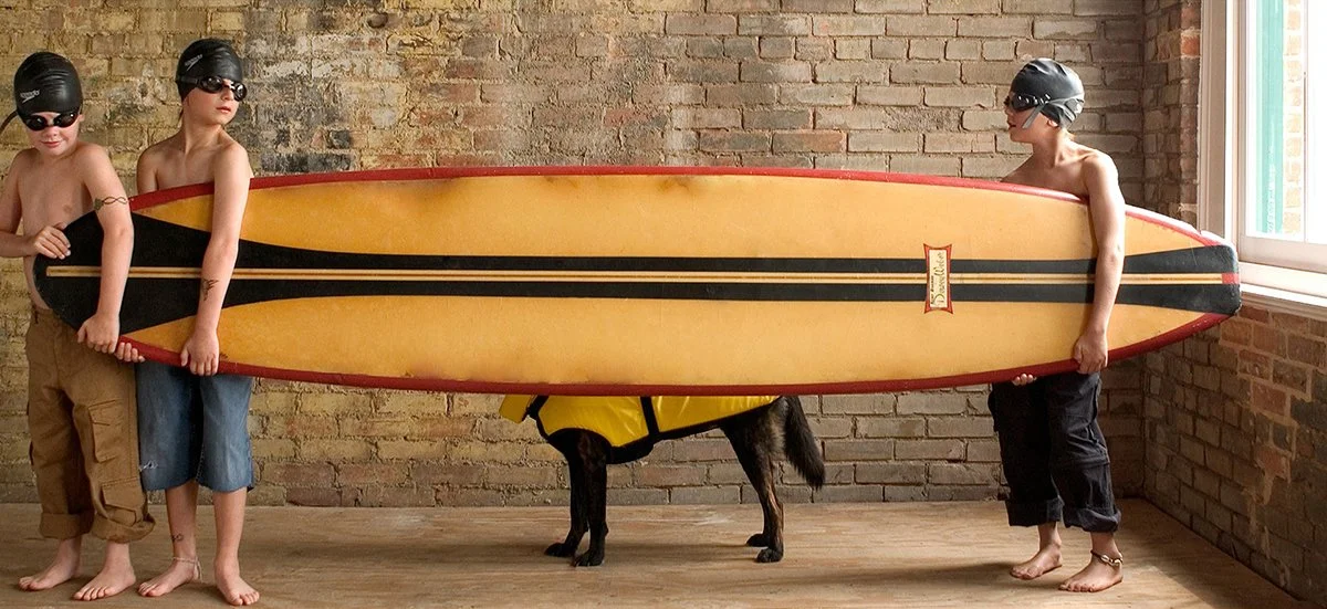 Three children holding a surfboard, wearing helmets and goggles, with a dog underneath it, in front of a brick wall.