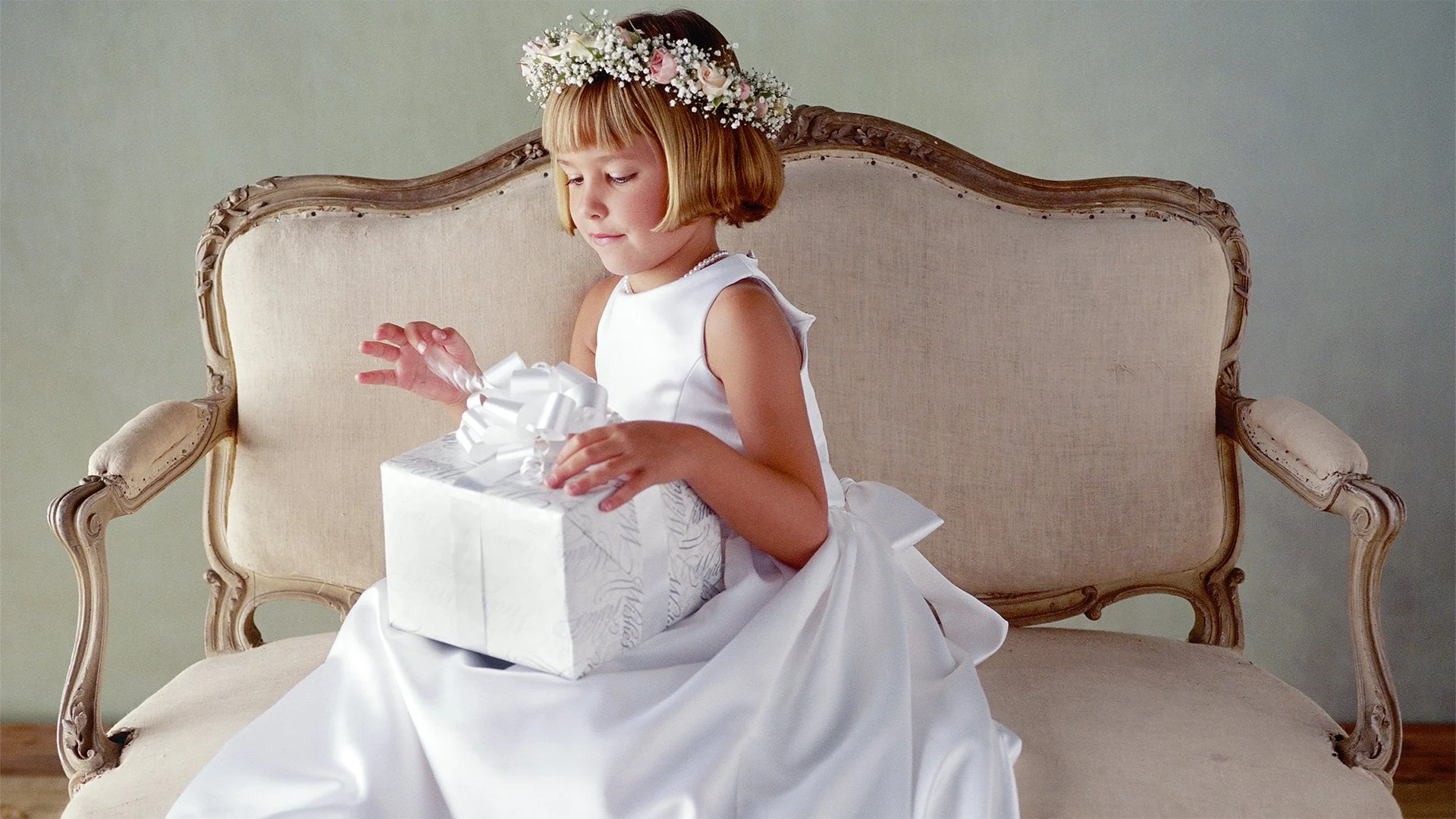 A young girl in a white dress with a floral headband sitting on a vintage-style sofa, holding a white gift box with a large bow.