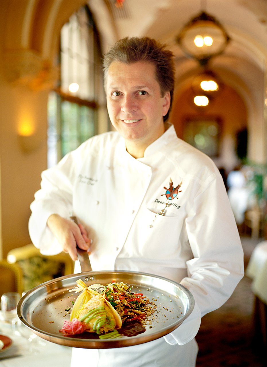 A chef in a white uniform holding a silver tray with a plated dish, smiling in a warmly lit restaurant with arched ceilings and hanging lights.