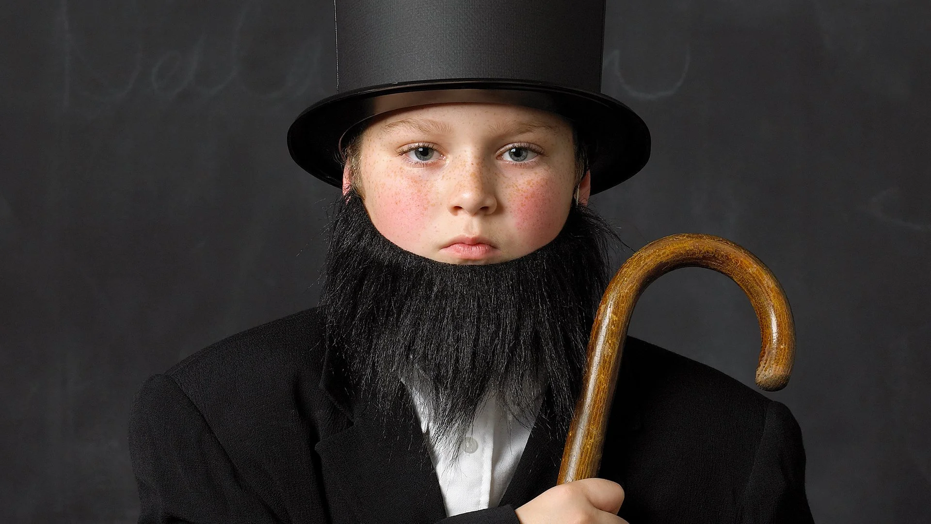 Young boy dressed as Abraham Lincoln wearing a black top hat, a fake black beard, and a black suit, holding a wooden cane against a dark background.