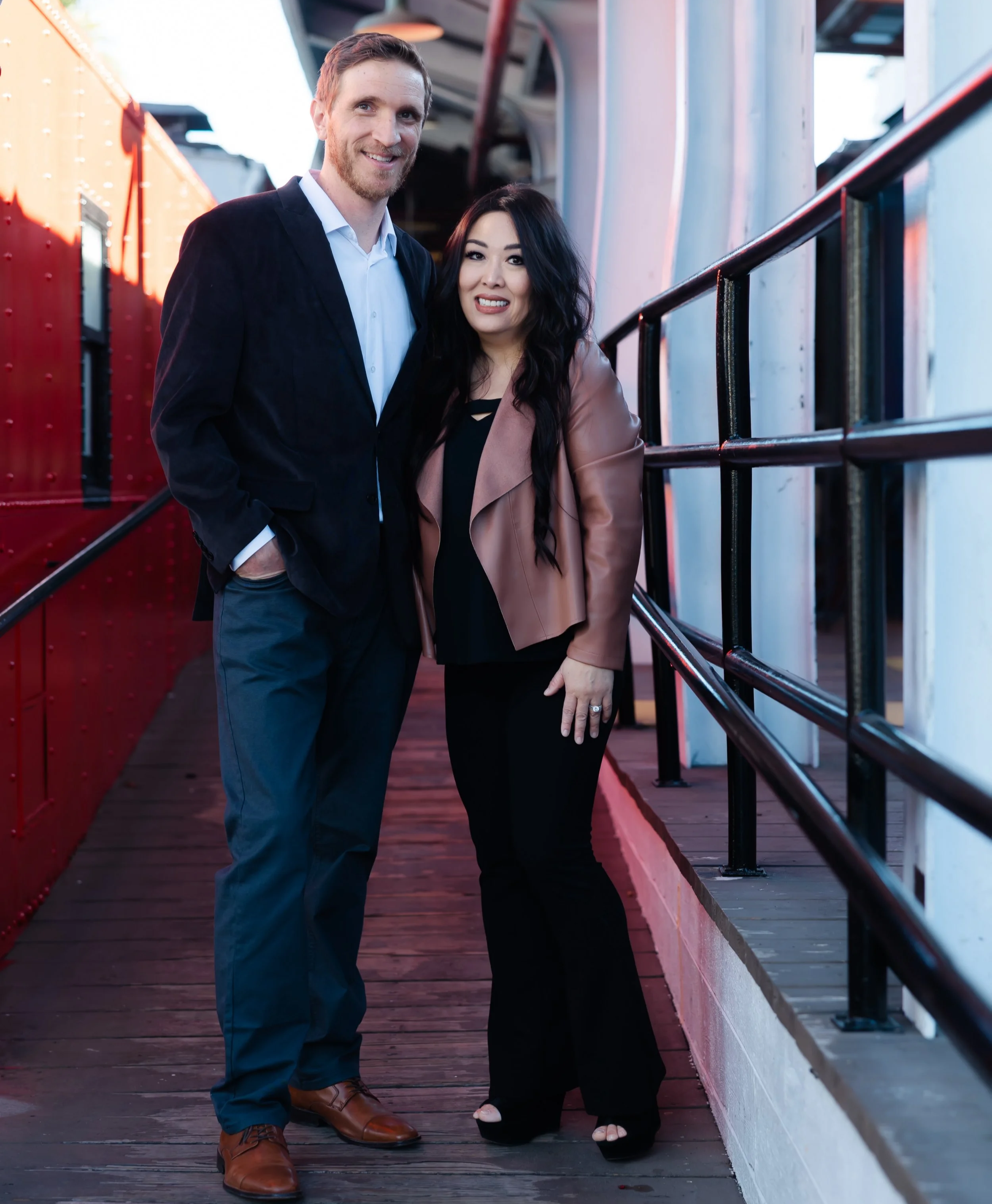 A man in a dark blazer and pants standing with a woman in black pants and a brown jacket with sheer sleeves on a wooden deck with a red and white structure in the background.