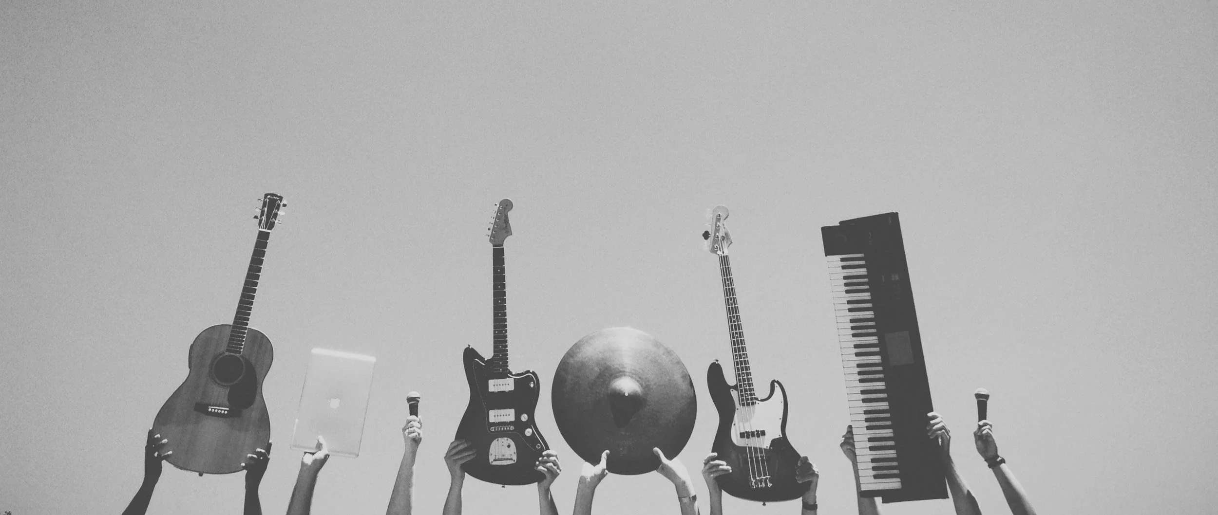 Multiple hands holding various musical instruments and microphones, including an acoustic guitar, electric guitar, cymbal, bass guitar, keyboard, and microphones, against a plain background.
