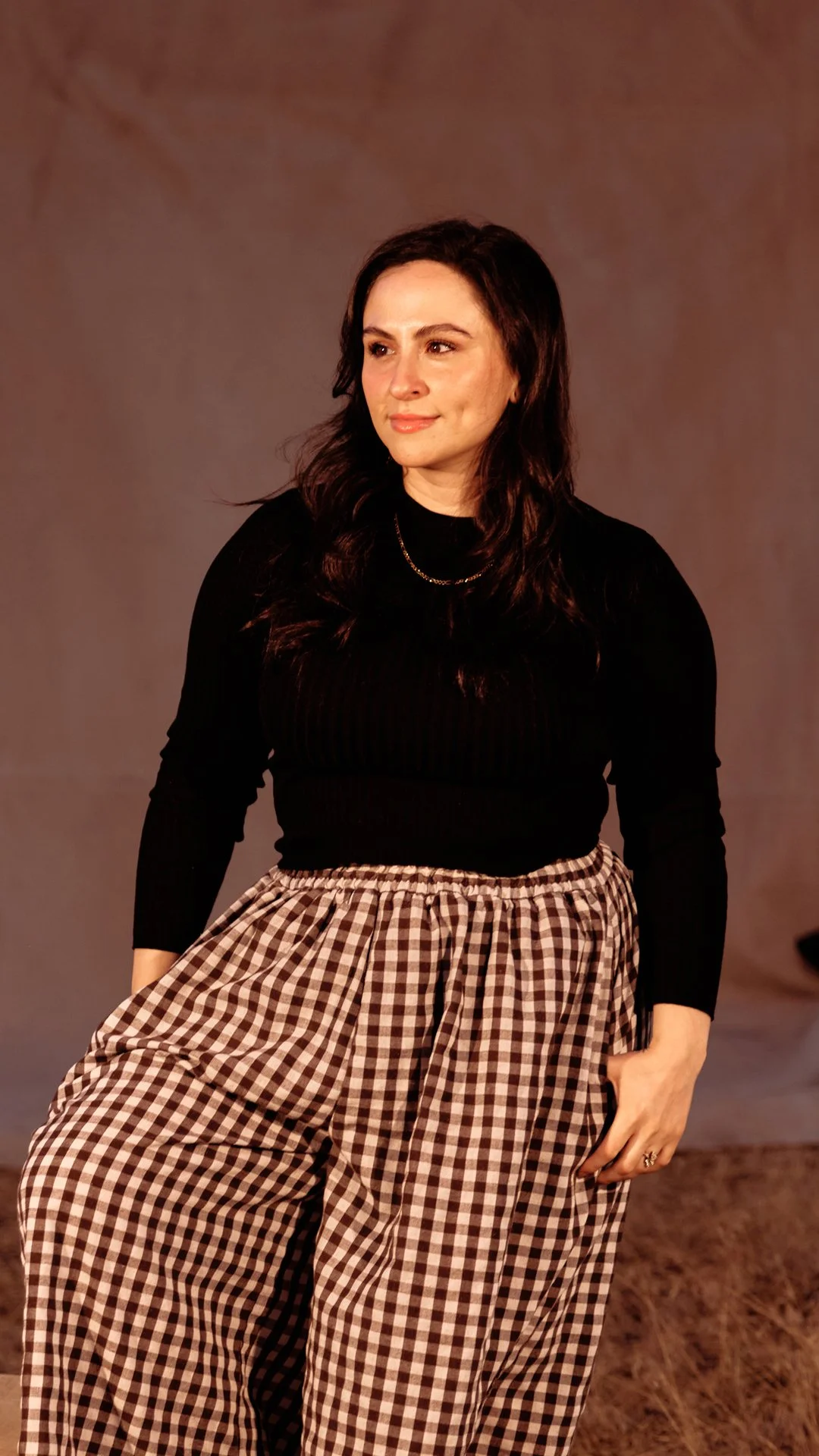 A woman with long, dark hair wearing a black long-sleeve top and brown and white checkered pants.