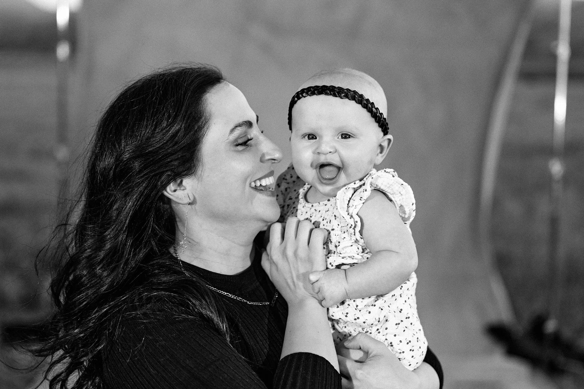 A woman with dark hair holding a smiling baby girl wearing a headband and floral dress.