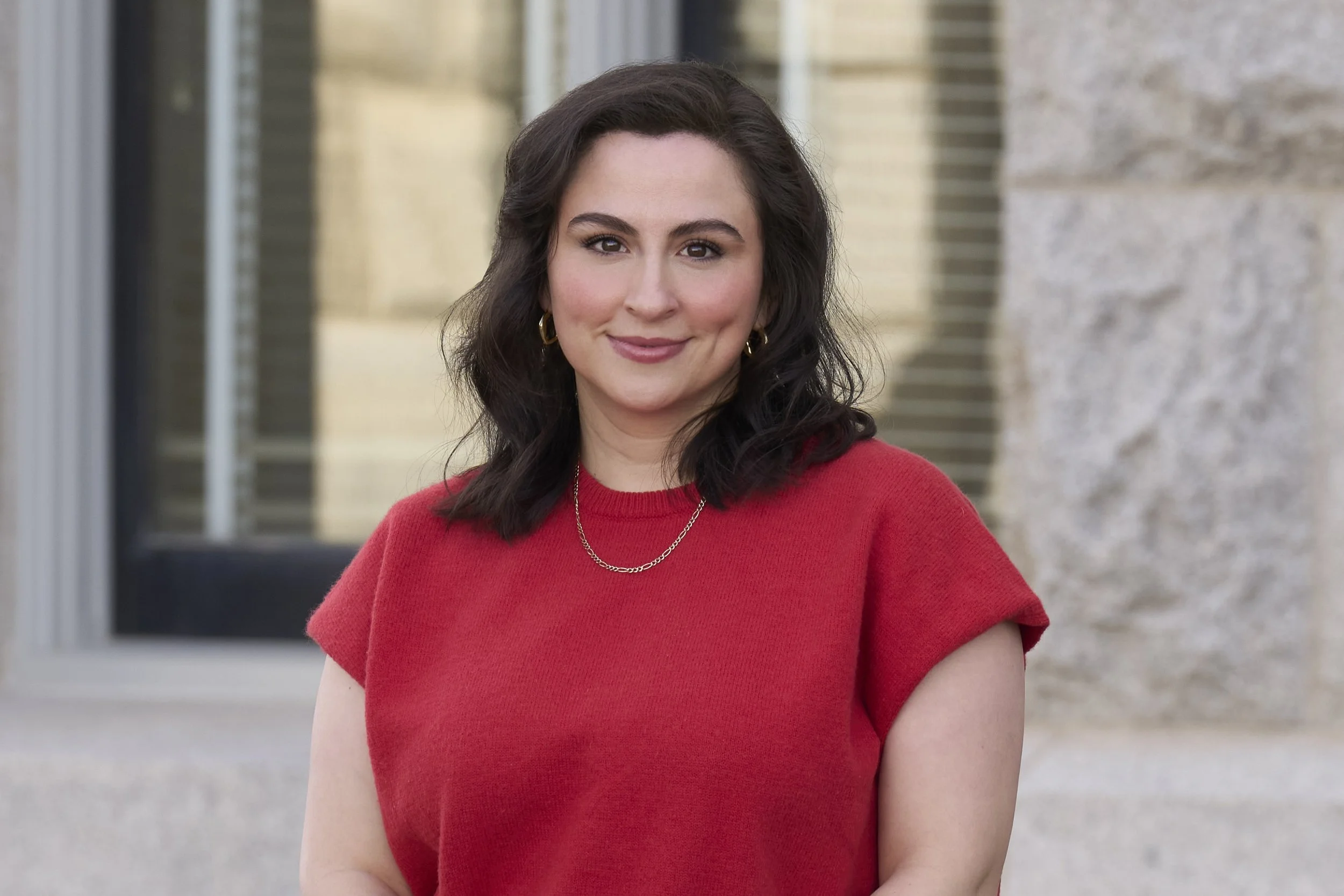 A woman with shoulder-length dark hair, wearing a red top, gold earrings, and a gold chain necklace, standing outdoors in front of a building with brick and stone walls.
