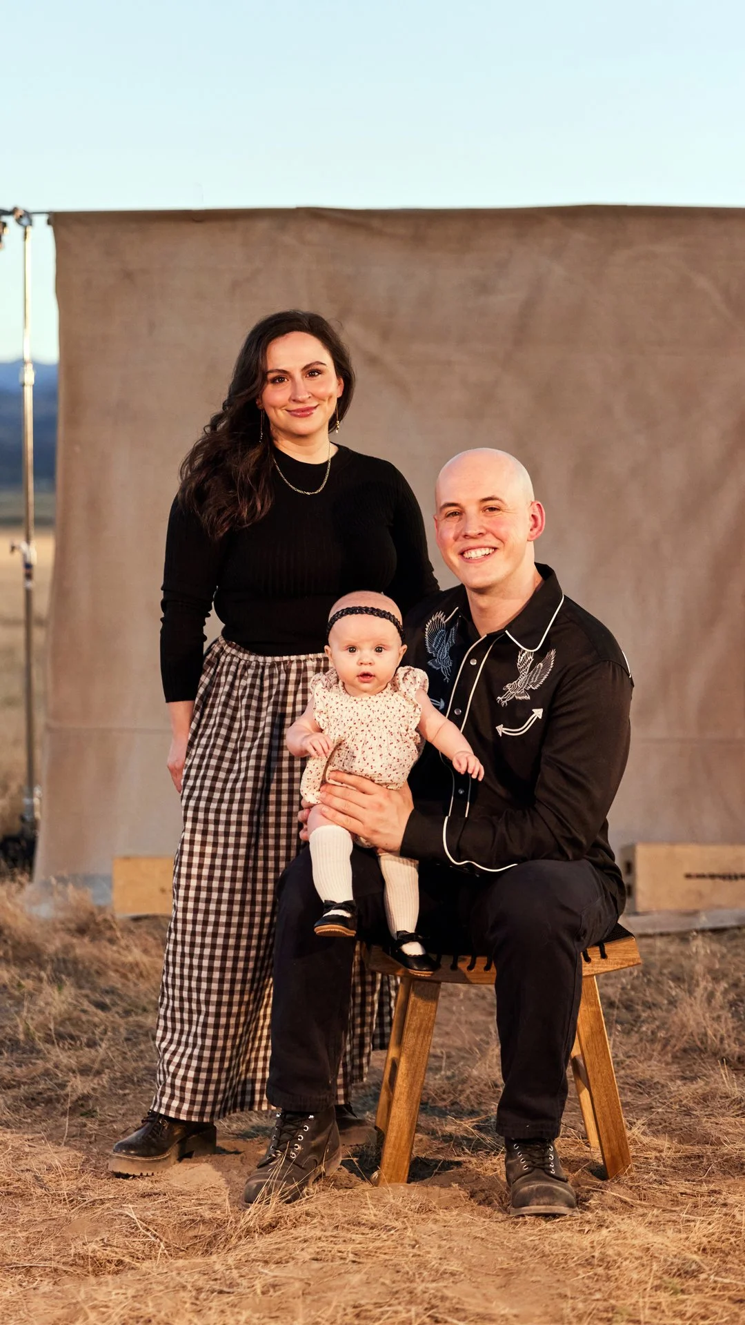 A family of three posing outdoors in front of a large backdrop. The woman is standing on the left, wearing a black top and checkered pants. The man, sitting on a stool, is holding a baby girl dressed in a floral outfit with a headband. The family is smiling at the camera.