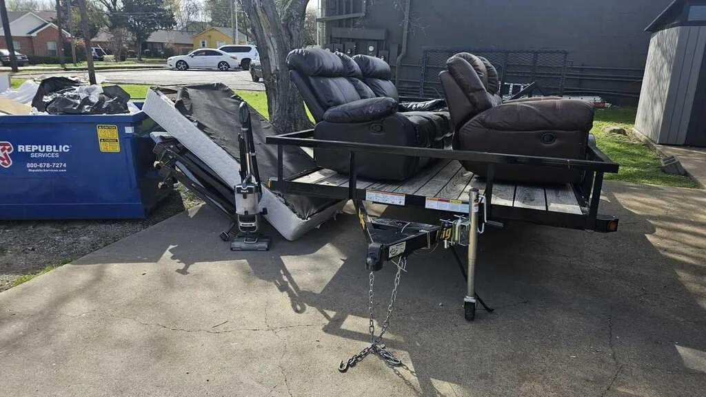 A trailer with two black recliner chairs and a dismantled black couch on a wooden platform, parked on a concrete driveway near a blue trash bin, with trees and houses in the background.