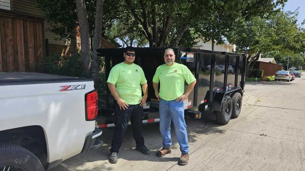 Two men in bright green shirts stand next to a black trailer hitched to a white pickup truck on a residential street.