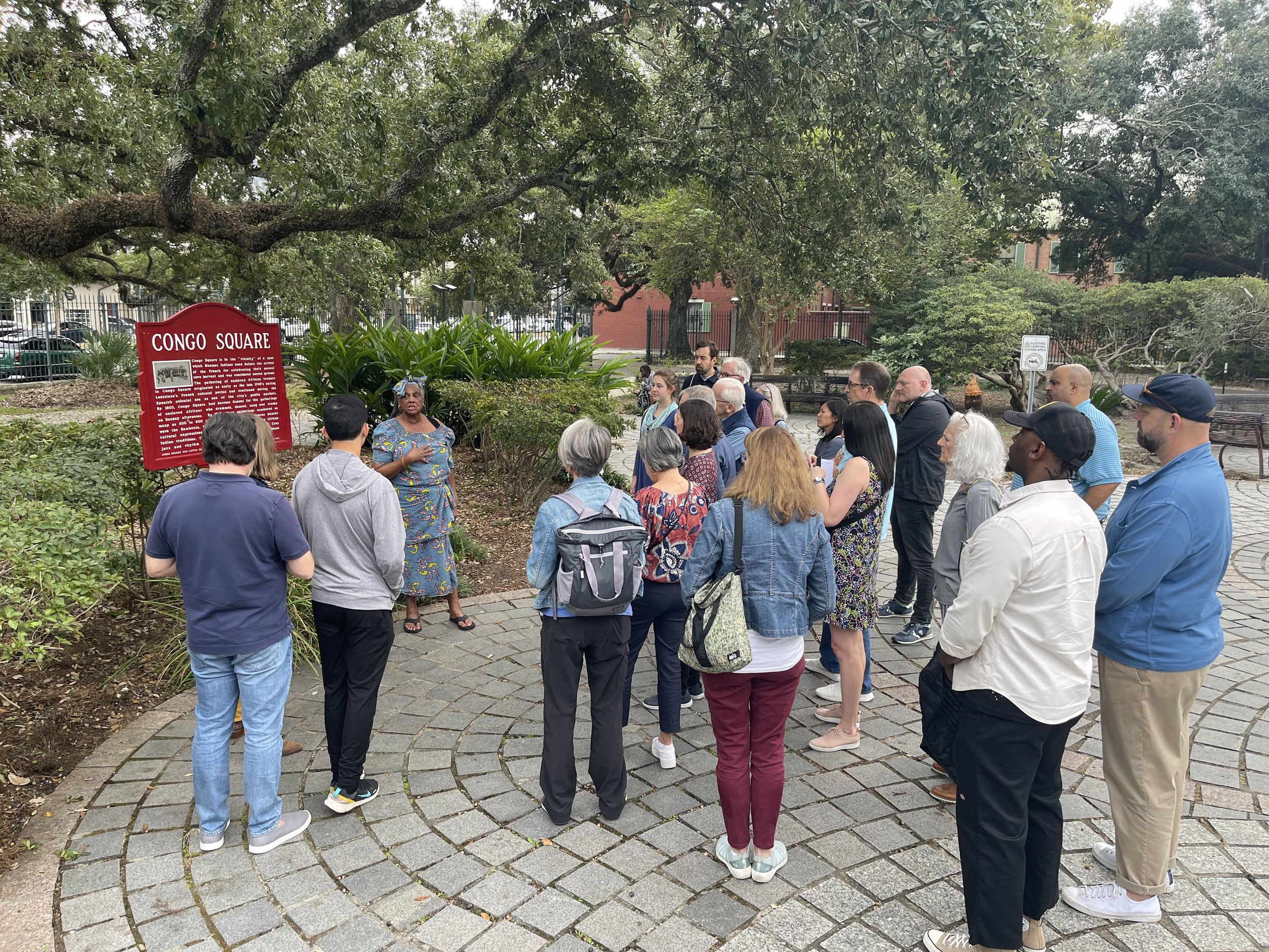 A diverse group of people gathered outdoors in a park, listening to a woman speaking. The scene features trees and greenery, with a red informational sign about Congo Square.