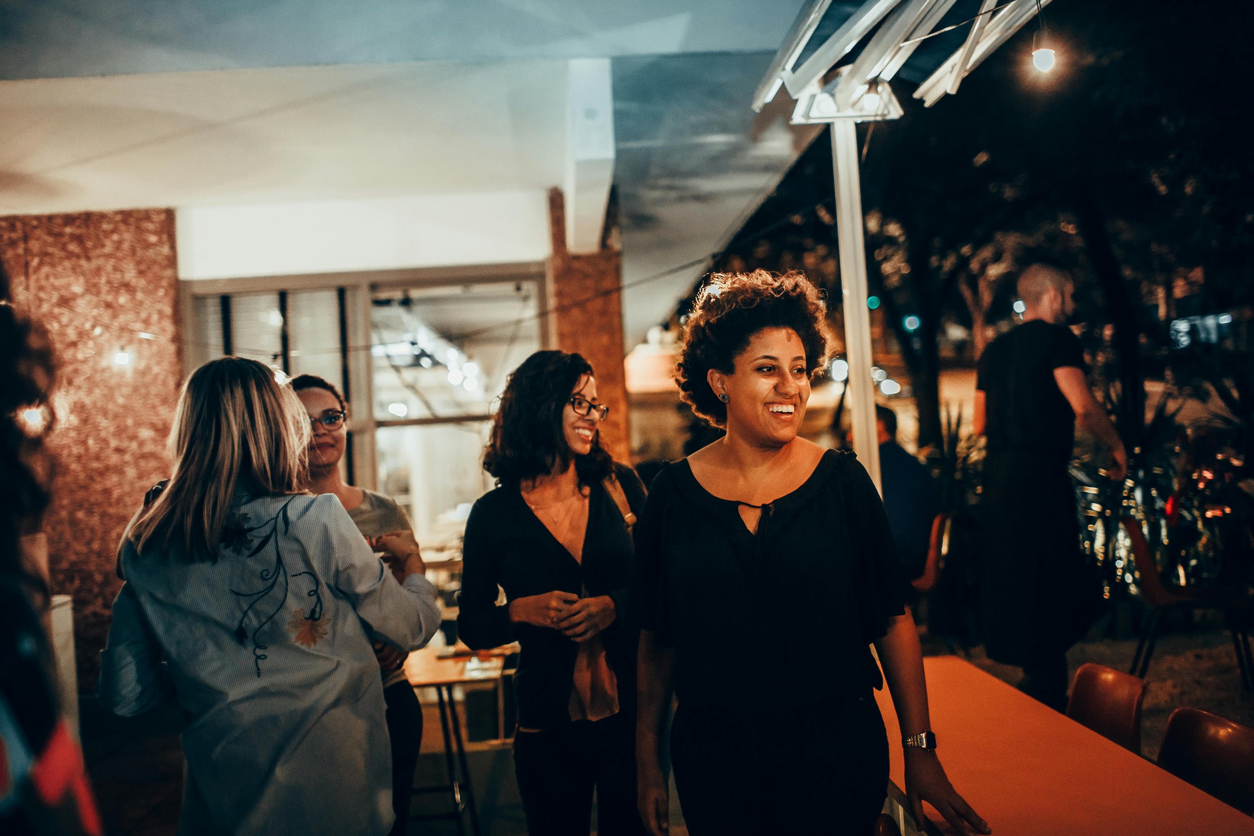 A group of smiling women socializing at an outdoor evening gathering, with string lights and tables in the background.