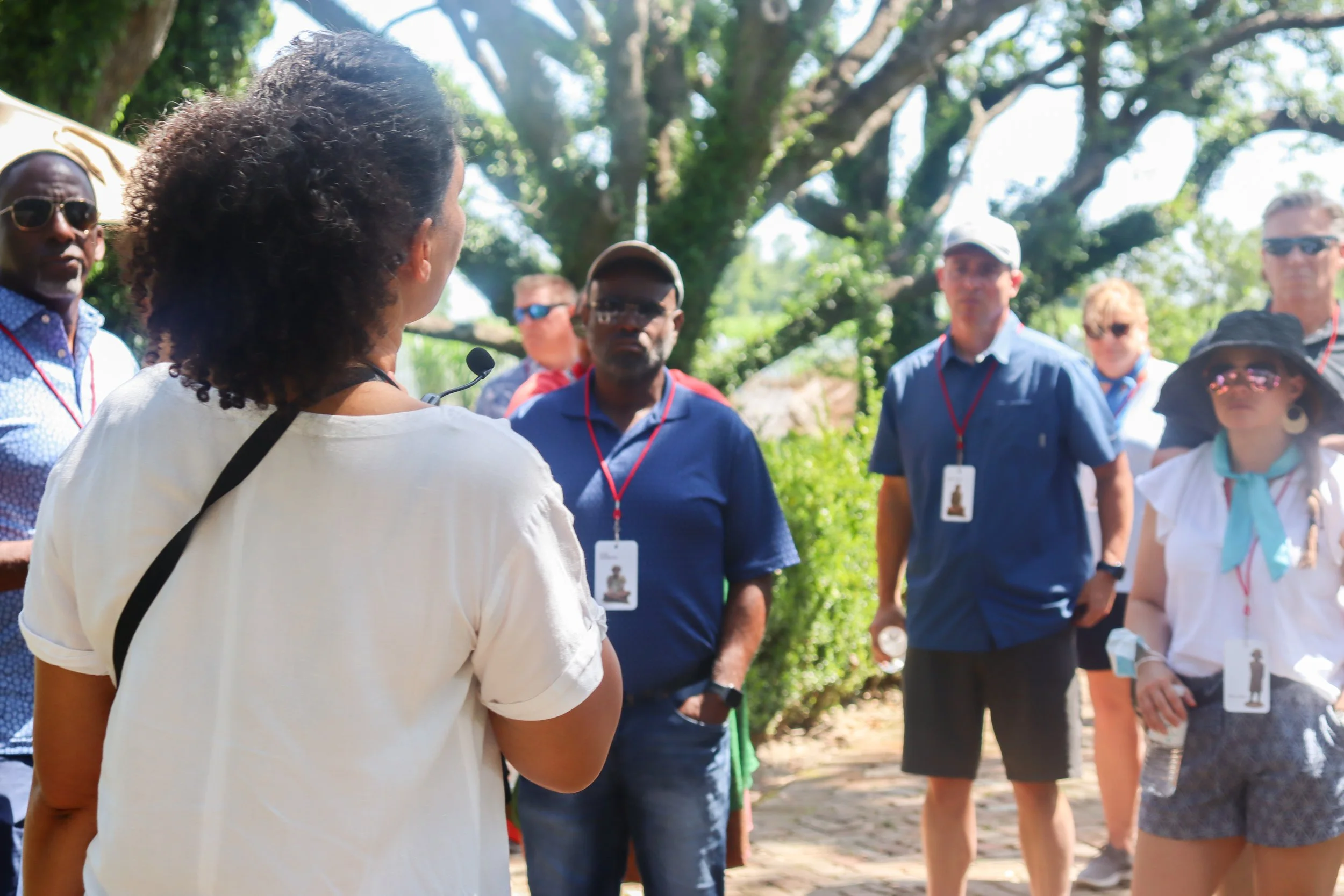 A woman with curly hair giving a talk to a group of people outdoors, with trees and sunlight in the background.