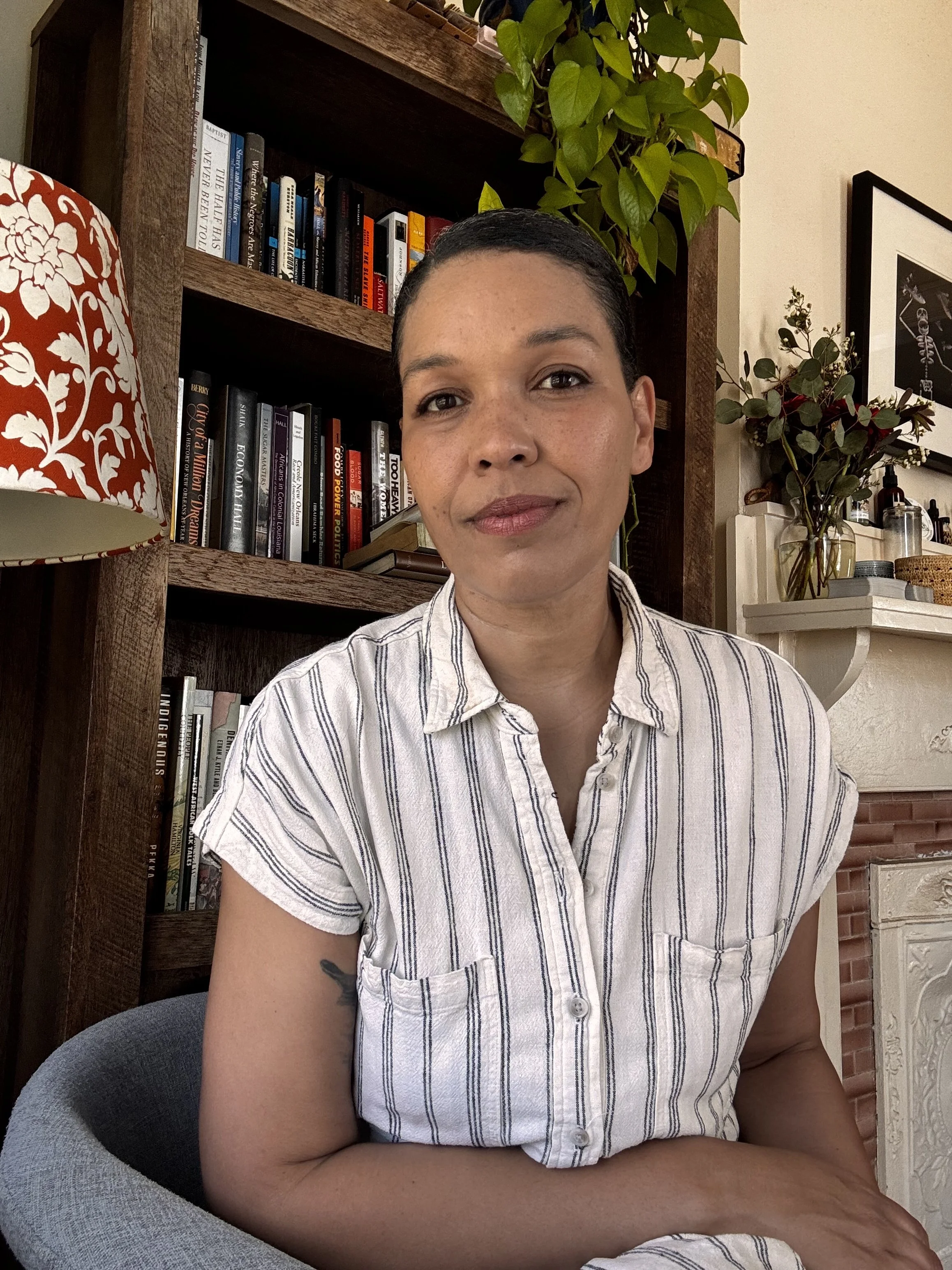 A woman with dark hair in a bun, wearing a striped shirt, sitting in a room with bookshelves and decorative plants behind her.