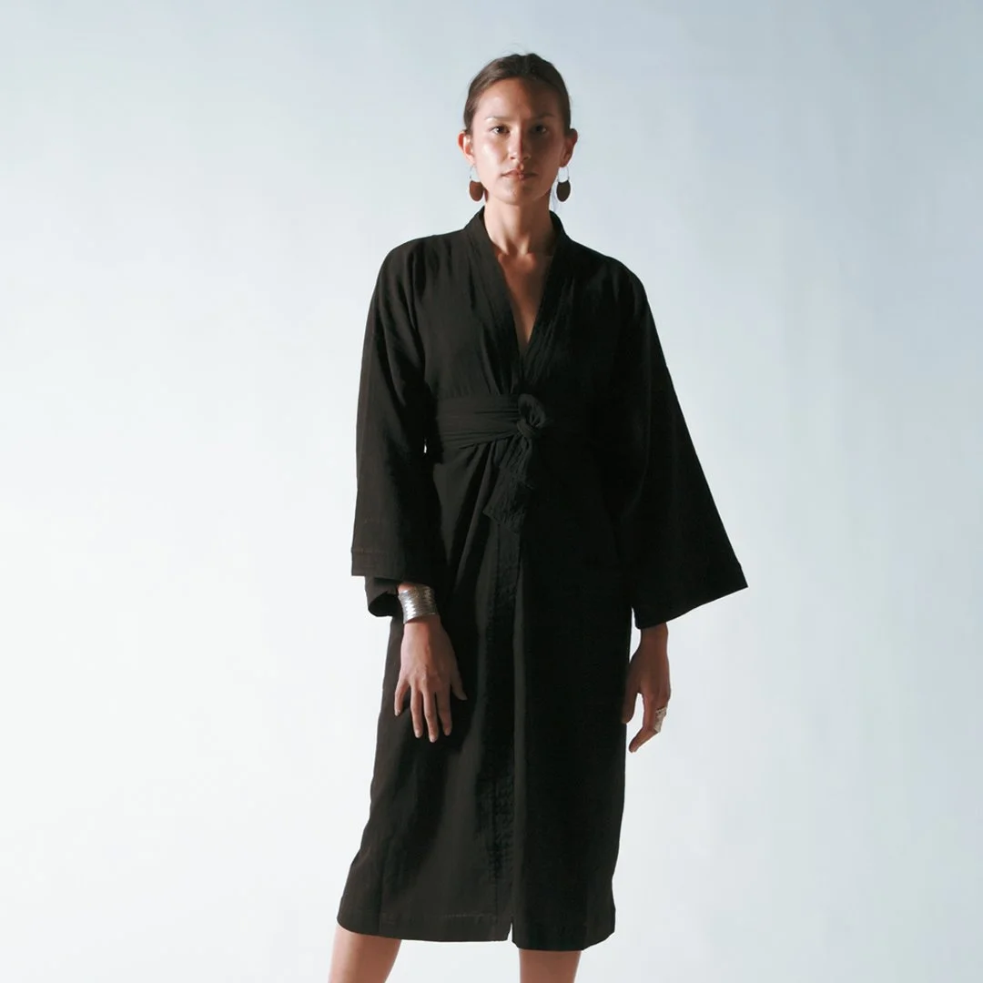 A woman with dark hair pulled back, wearing a long black dress with wide sleeves and a tied waist, standing against a plain light background.