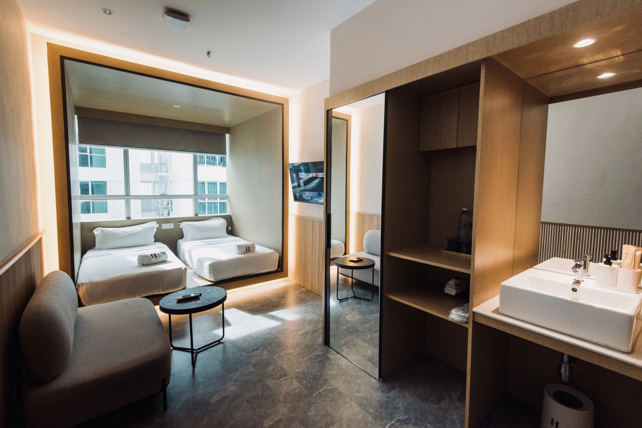 Modern hotel room with two twin beds, a small sofa, a round coffee table, a flat-screen TV, a mirror, a chair, a bathroom sink with toiletries, and a large window with city view.