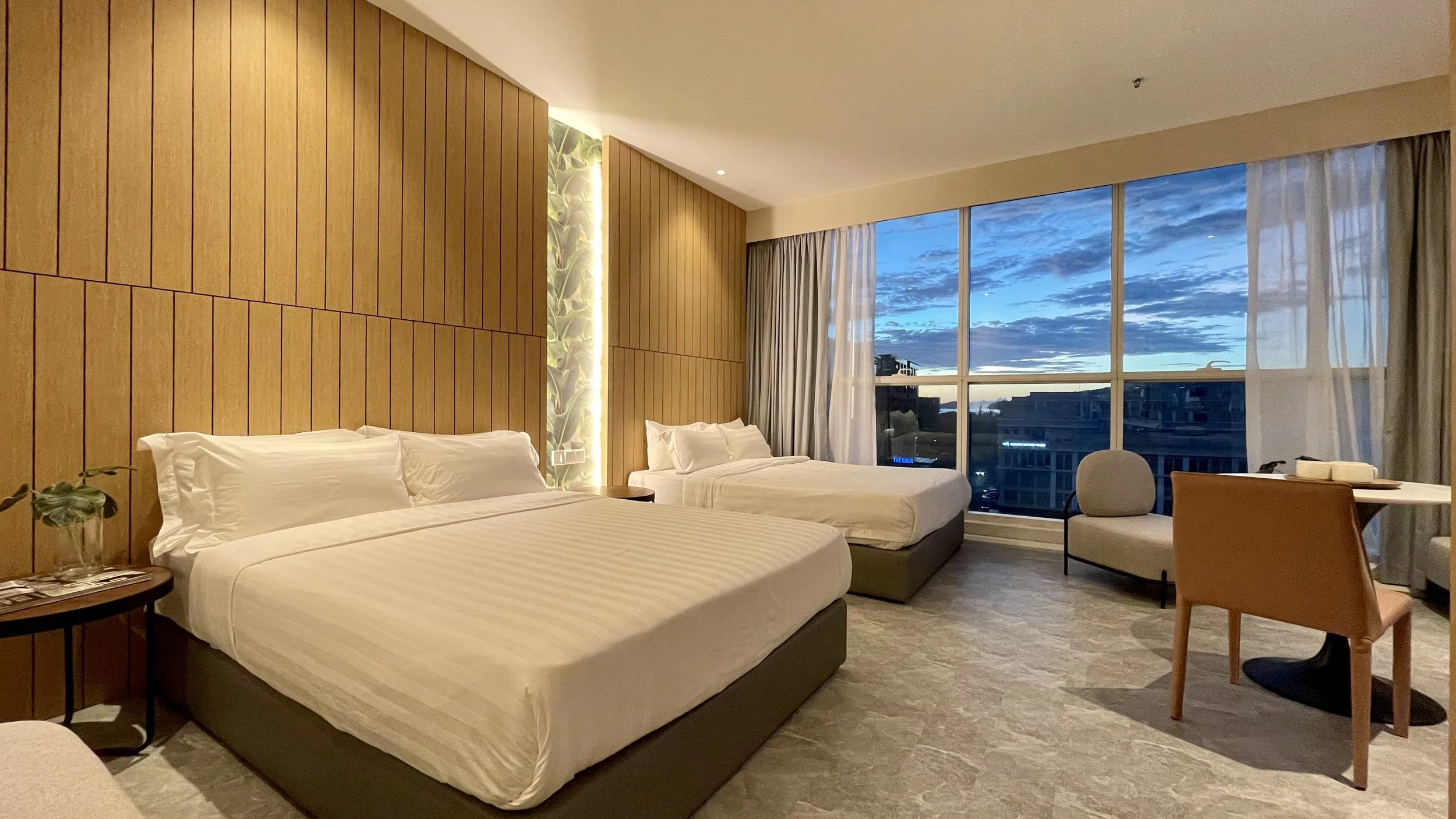 Hotel room with two beds, a large window showing a cityscape at dusk, and modern decor including wooden wall panels and a small seating area.