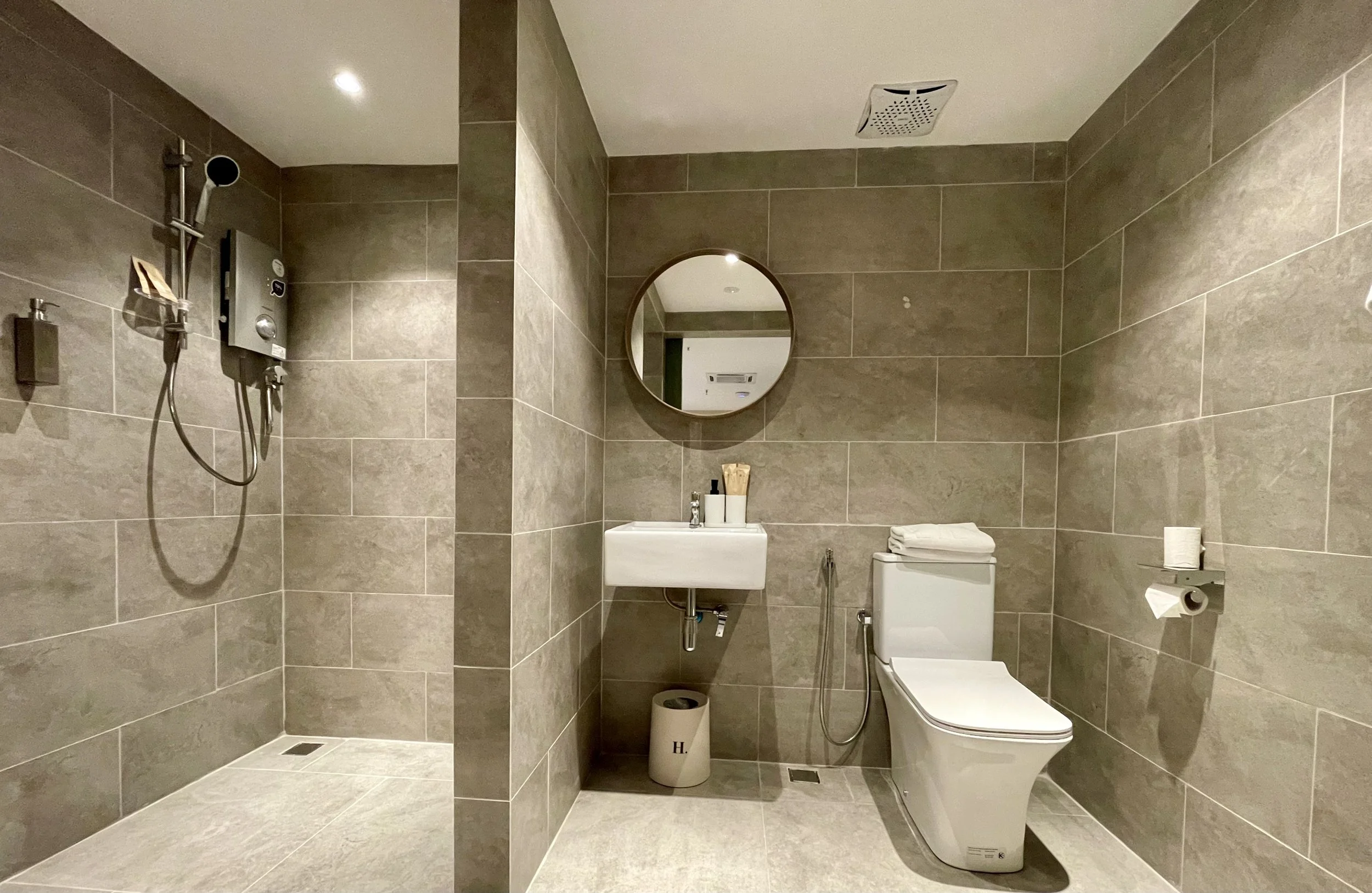 Modern bathroom with gray tiles, a walk-in shower area on the left, a white toilet on the right, and a small white sink with a round mirror above. There are towels and toiletries on the sink and a toilet paper roll on the wall.