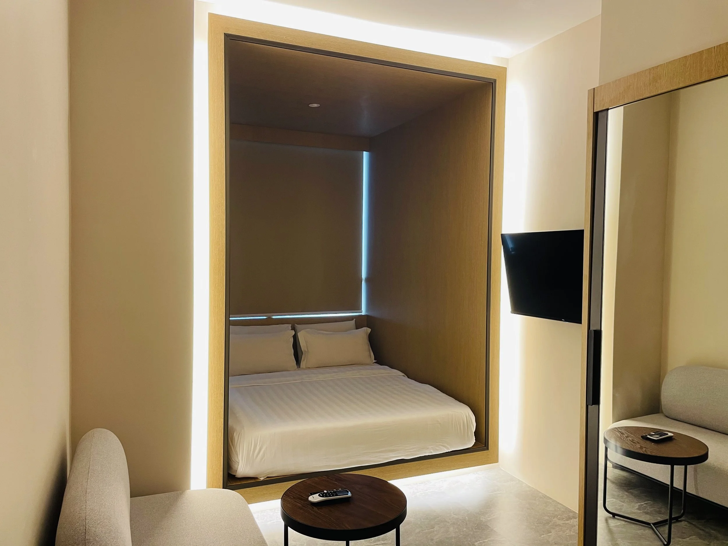 Modern hotel room with a bed inside a wooden framed alcove, a wall-mounted TV, and a seating area with a round side table and remote control.