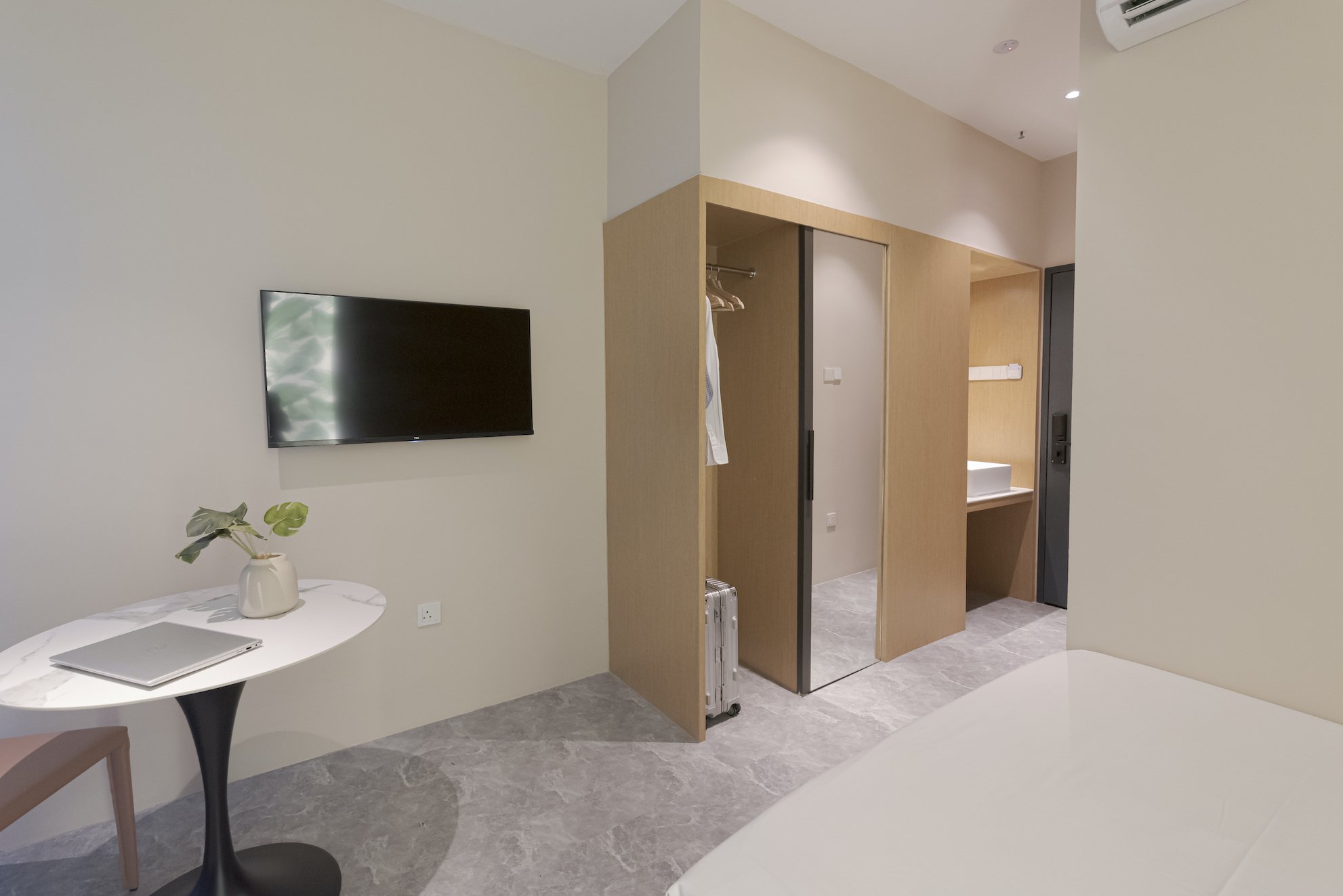 Interior of a modern hotel room with a wall-mounted TV, a small table with a laptop and a plant, a closet with hanging clothes, and a bathroom counter with a sink.