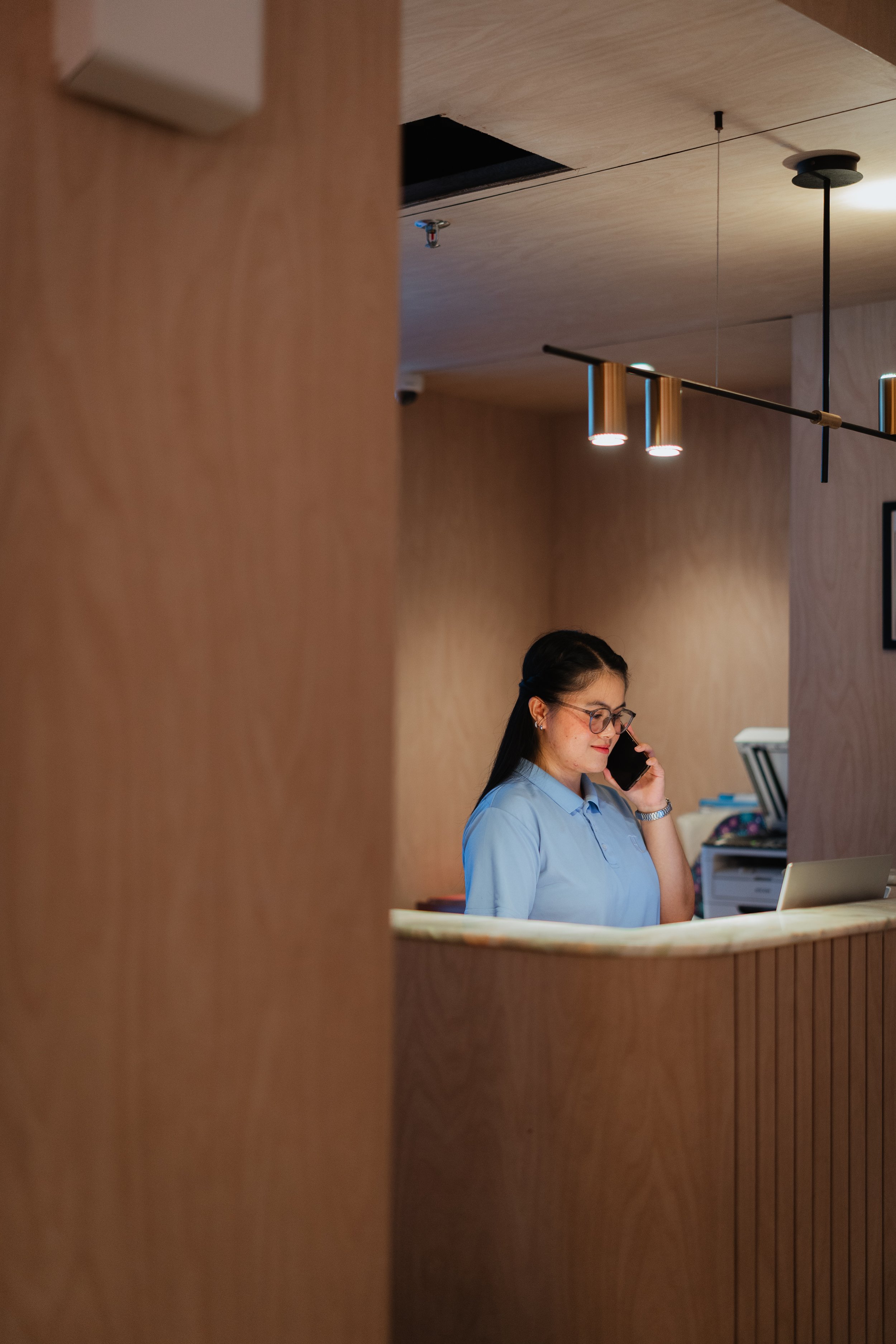 A woman wearing glasses and a light blue shirt speaking on a cellphone behind a wooden reception desk.