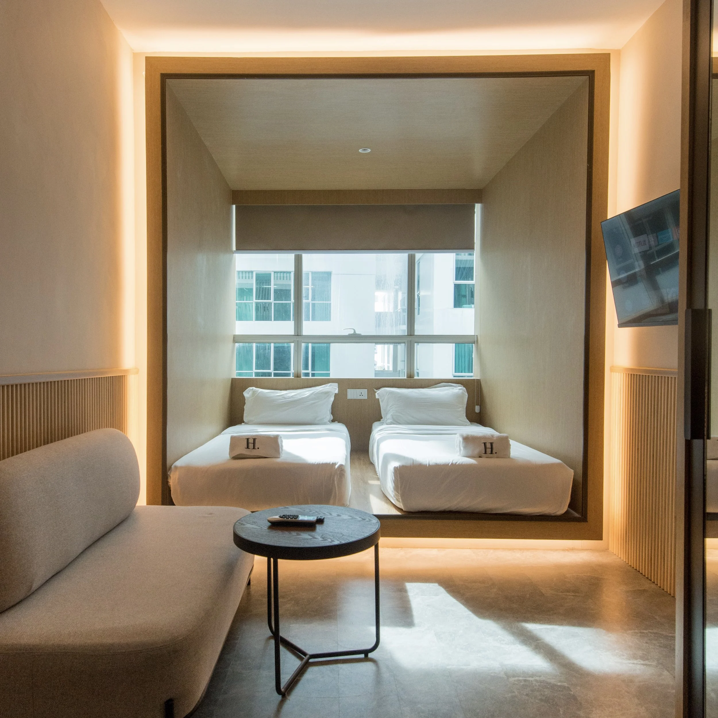 Modern hotel room with two single beds, a large window, a beige sofa, a round black side table, and a wall-mounted TV.