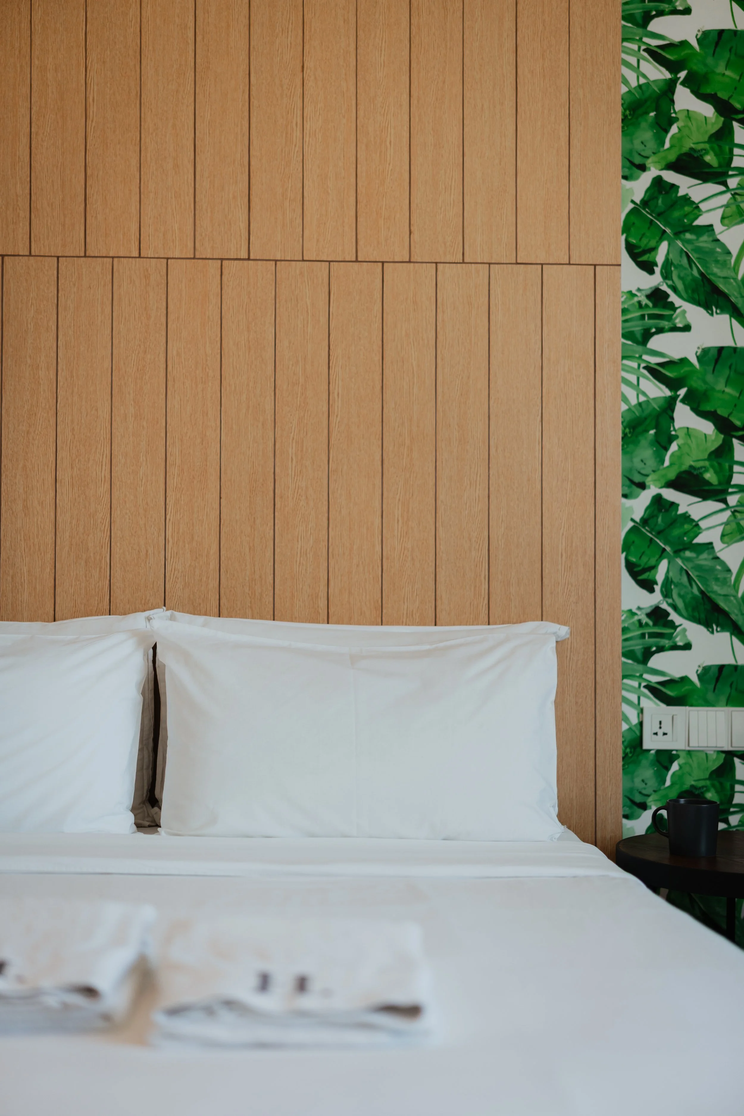 A neatly made bed with white pillows and sheets, a black mug on a black side table, a wooden headboard, and tropical green leaf wallpaper on the wall behind.