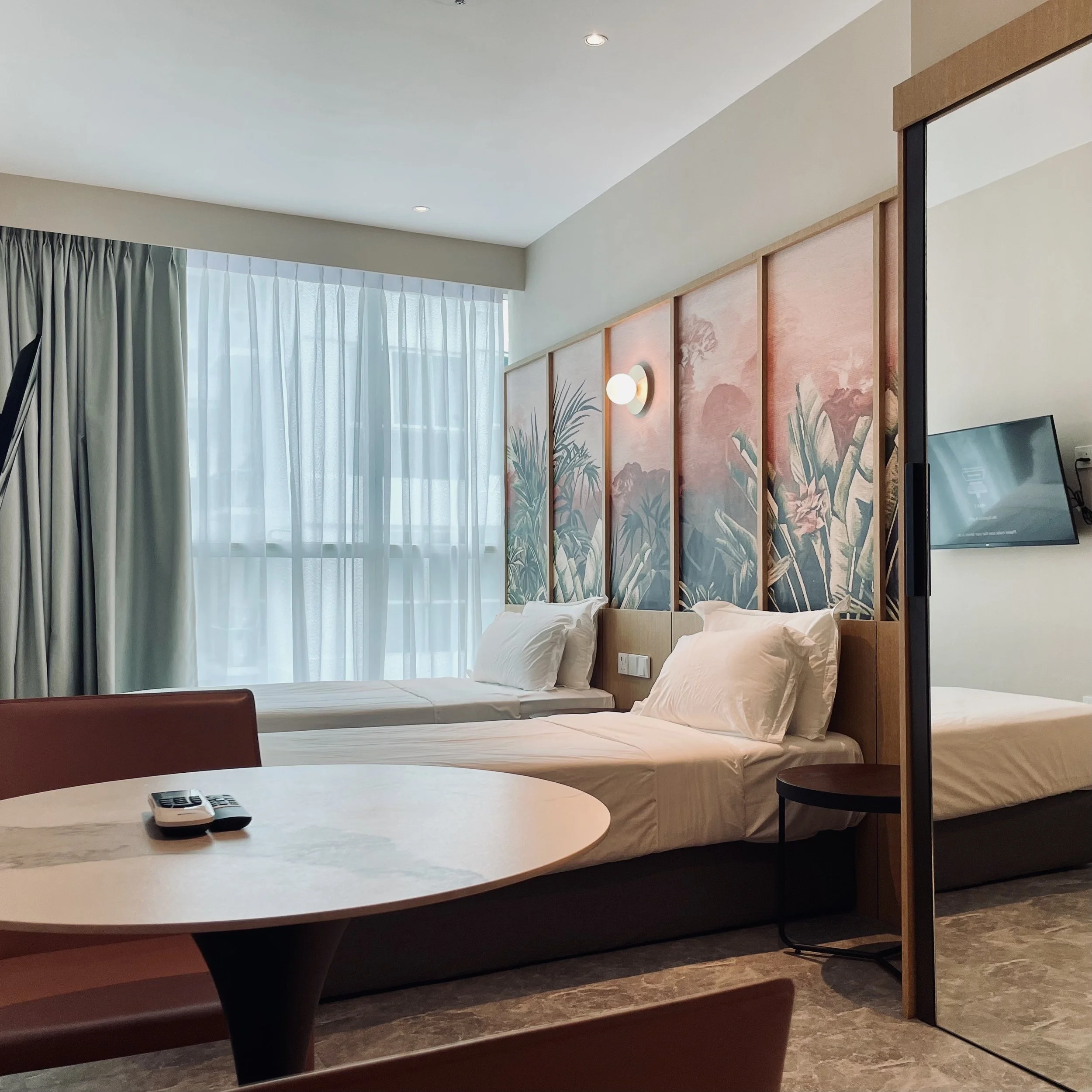 Modern hotel room with two single beds, a large window with sheer curtains, a round table with remote controls, wall art with botanical designs, a wall-mounted TV, and a small bedside table.