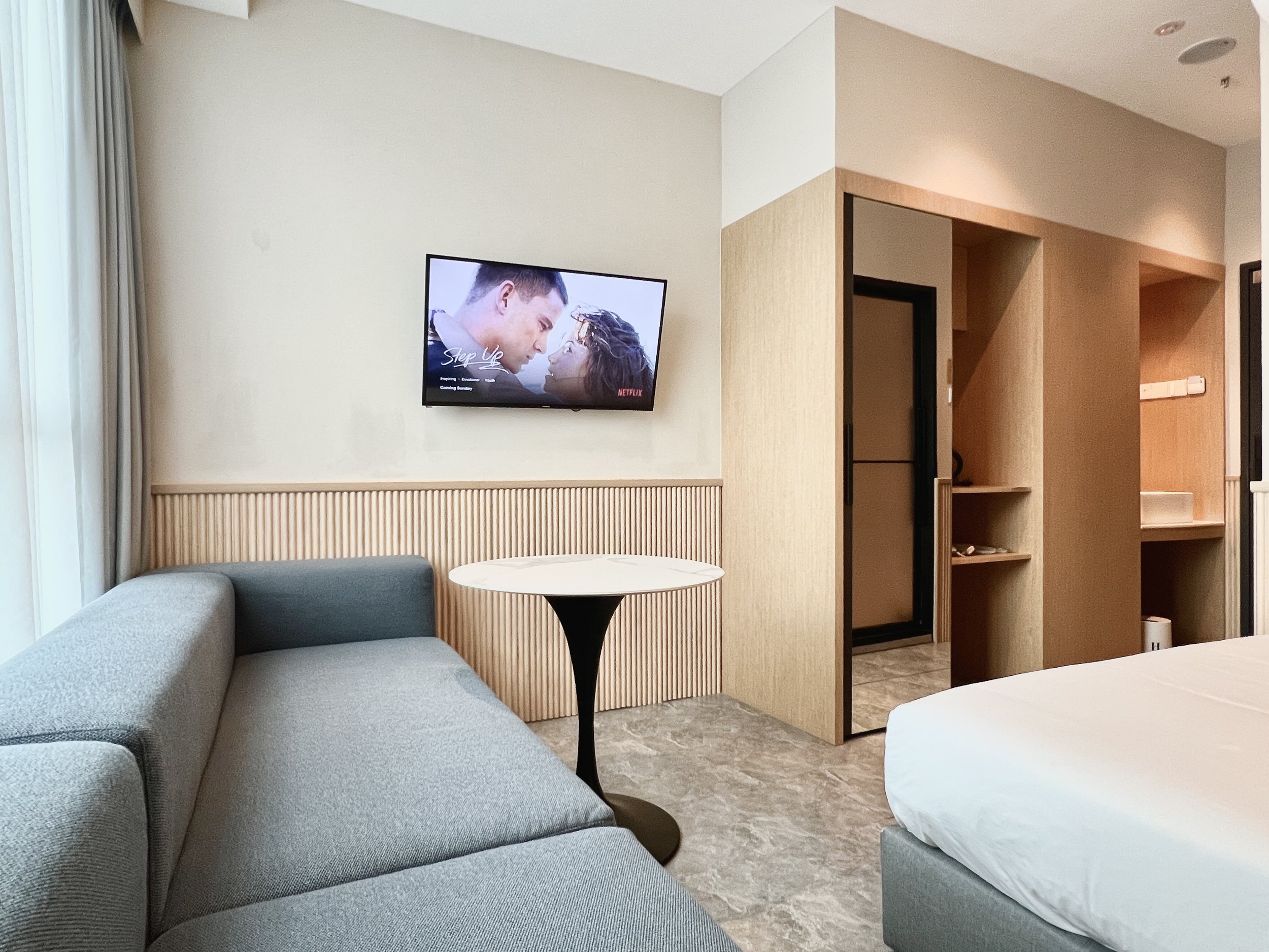 A modern hotel room with a gray sofa, a small round white table, a wall-mounted flat-screen TV displaying a romantic scene, and a partially visible bed on the right. The room has a minimalist design with light wood accents and stone flooring.