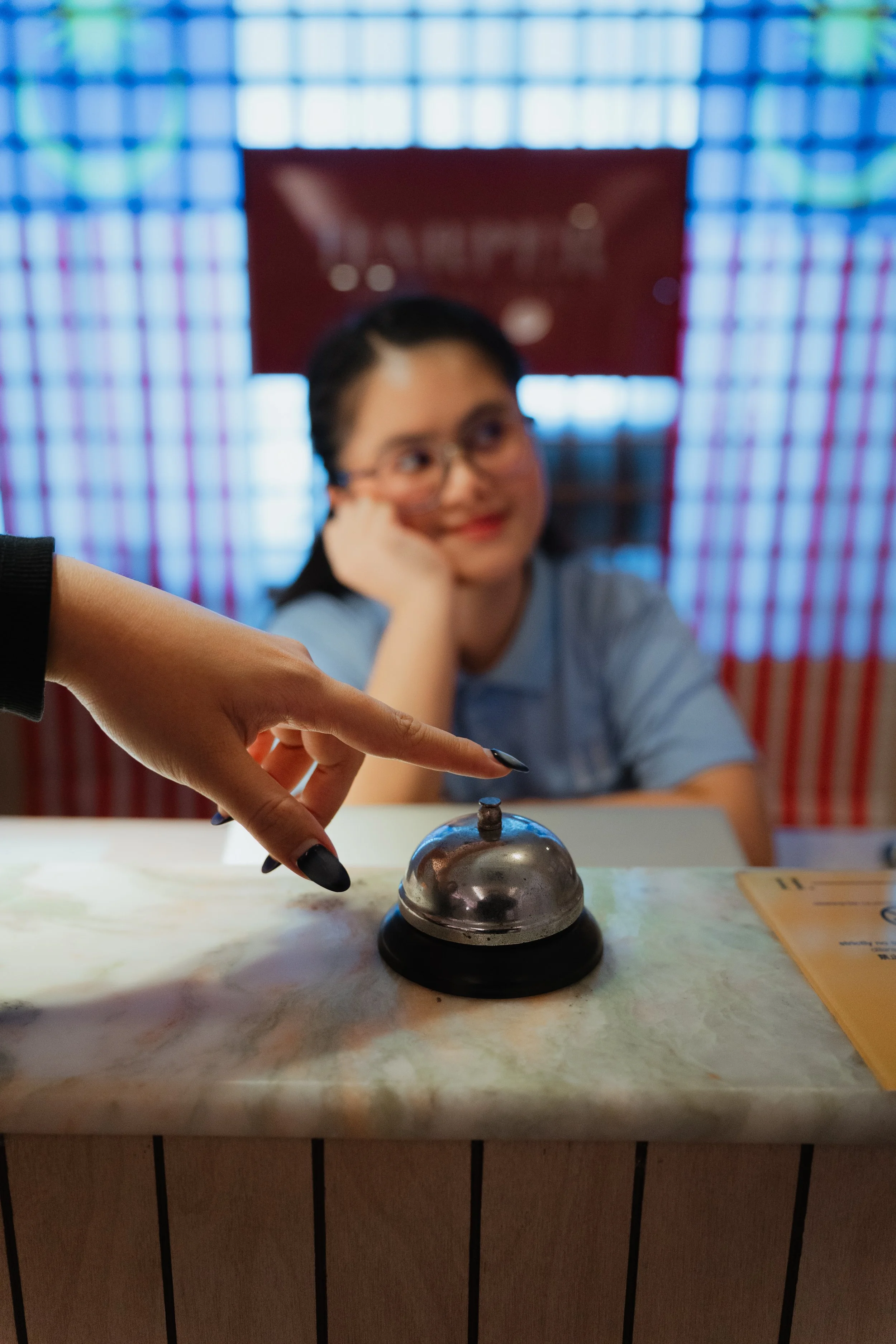 Person with black nail polish strikes a bell with an index finger at a reception desk, in front of a woman wearing glasses and a light blue shirt, sitting with her head resting on her hand, in a setting with colorful checkered backdrop.