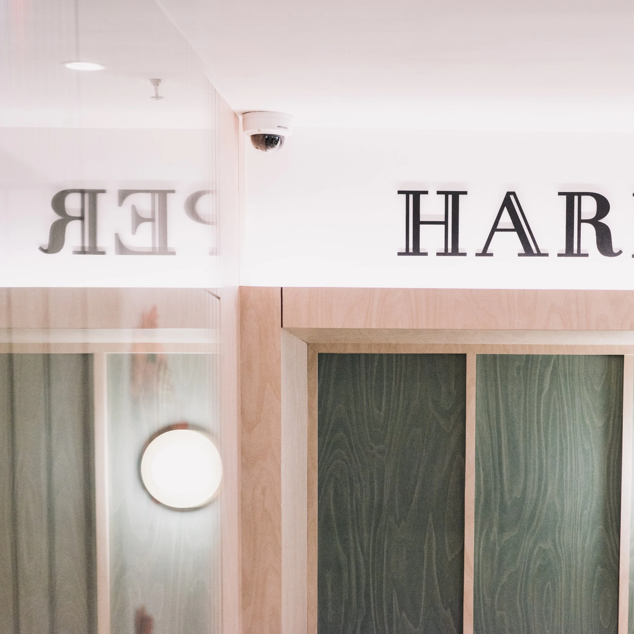 The image shows the reflection of a sign in a mirror, with the partial text 'HARD' visible. There is a wall-mounted security camera and a round ceiling light in the scene.