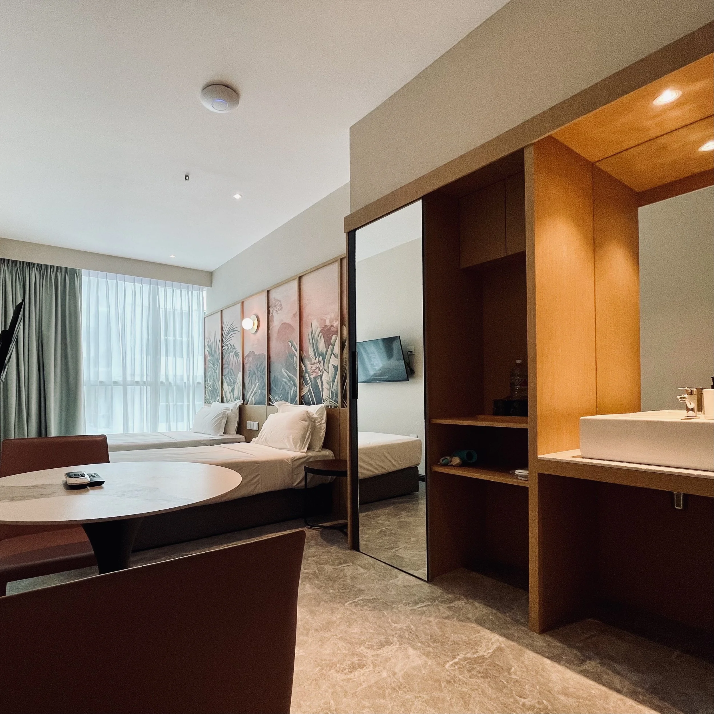 A hotel room with two beds, a round dining table and chairs, large window with curtains, decorative wall panels, a flat-screen TV, and a bathroom sink area with wooden cabinetry.
