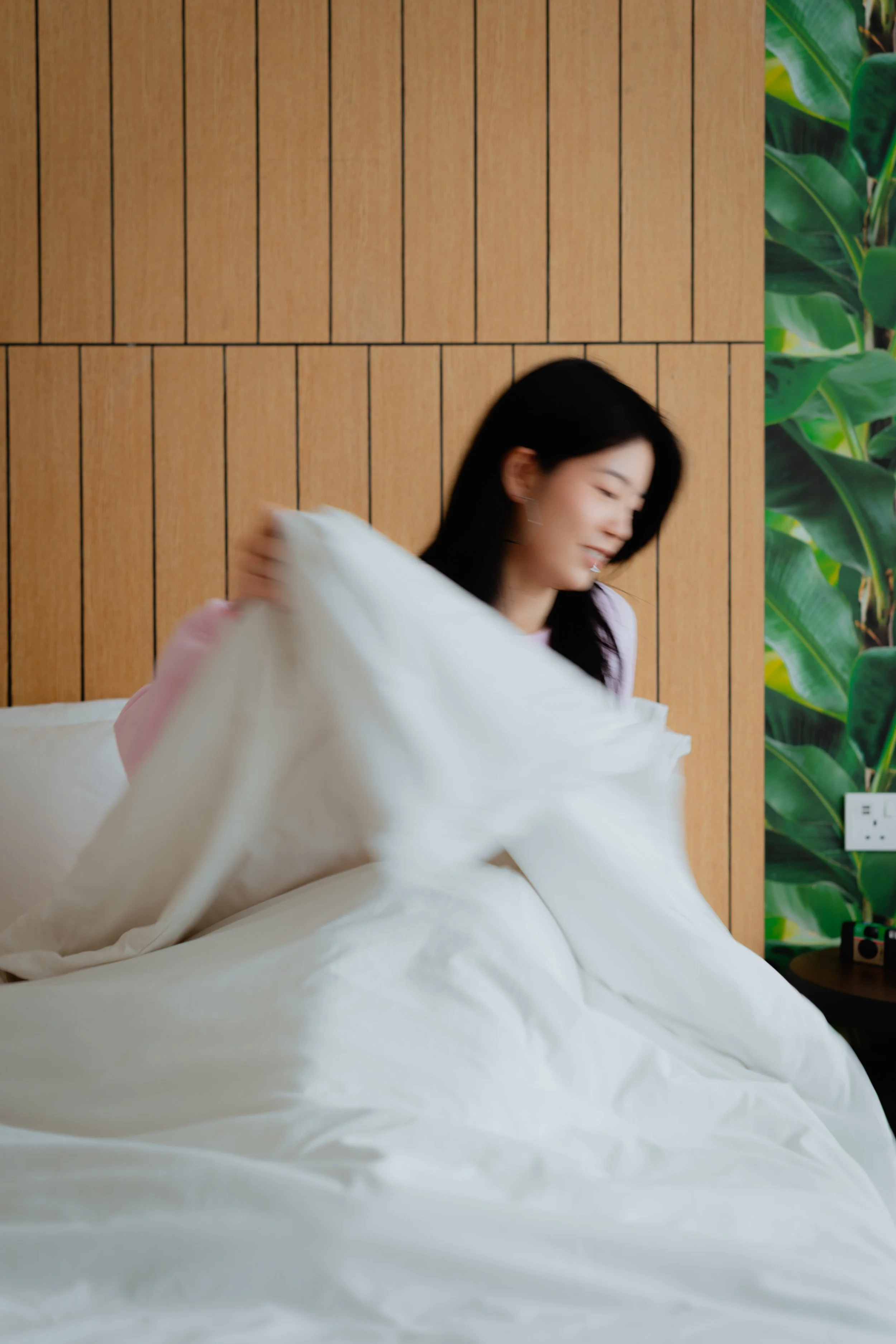 Woman making the bed with white sheets in a modern bedroom with wooden panel wall and green plant wallpaper.