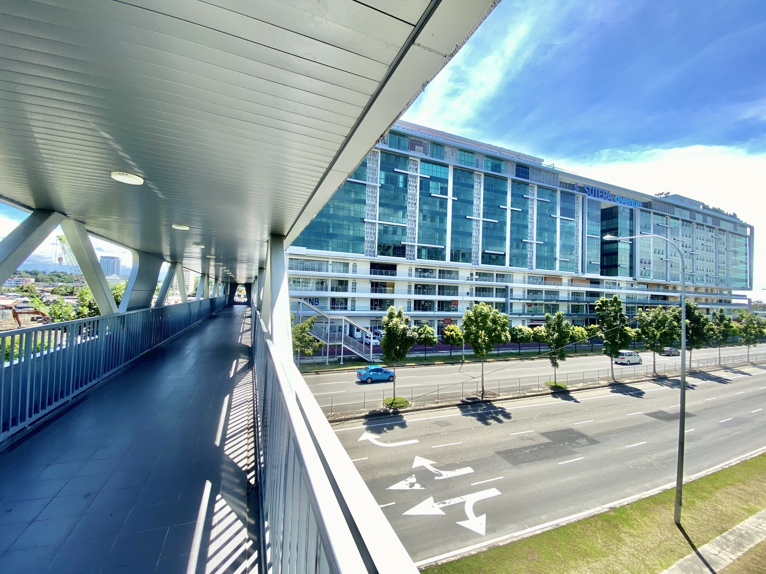View from an elevated walkway in an urban area showing a multi-road street with cars, a row of small trees, and a modern glass building labeled 'SUTERA Avenue' in the background, under a partly cloudy sky.