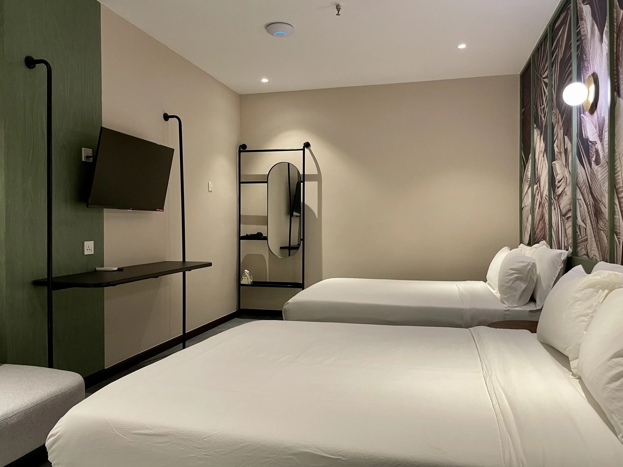 A hotel room with two white beds, a wall-mounted flat-screen TV, a small green accent wall, a modern black metal-frame mirror and shelf unit, and a decorative purple and green leaf patterned wall panel behind the beds.