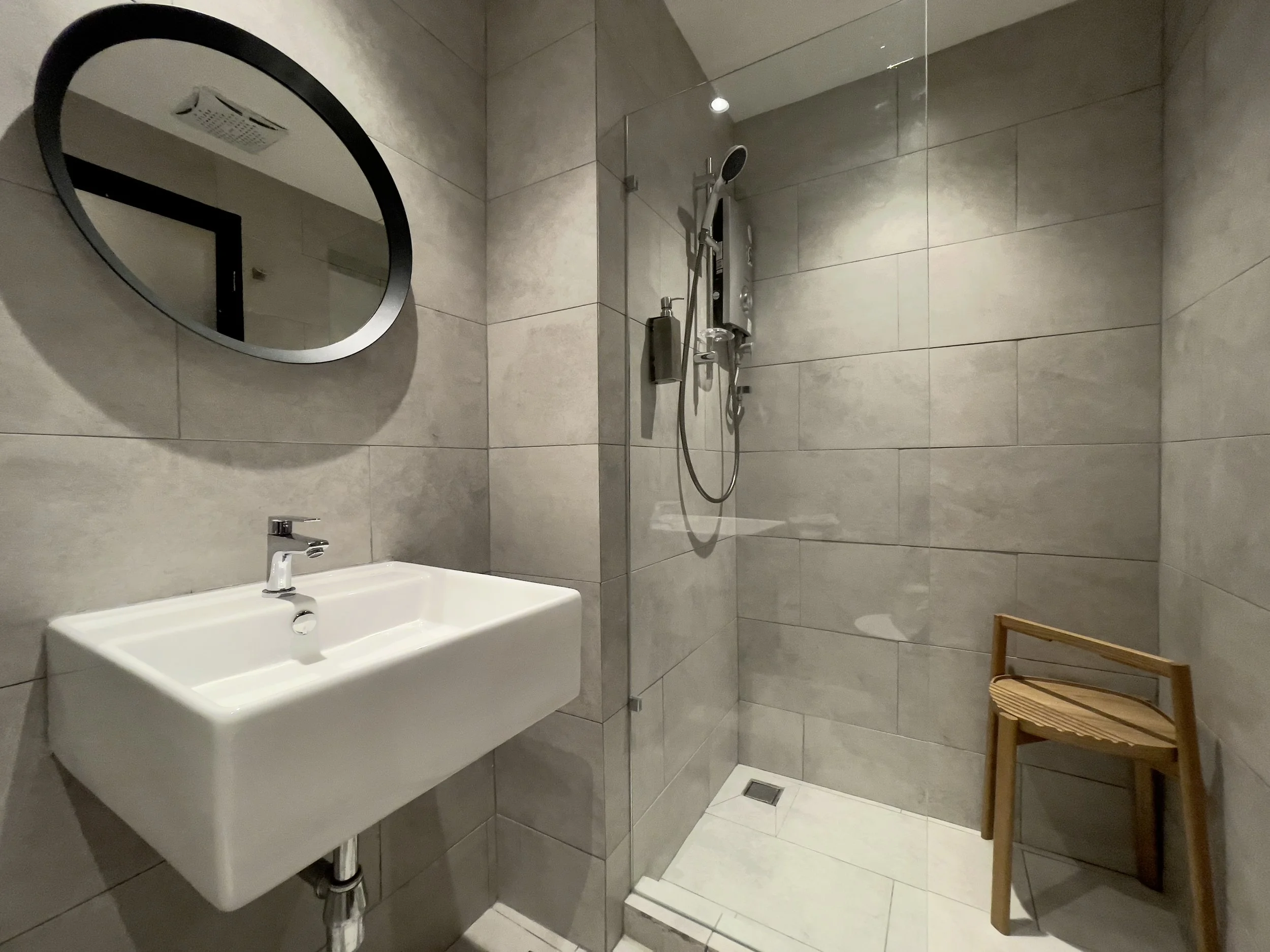 A modern bathroom with a wall-mounted rectangular white sink, a round mirror, and a walk-in shower with grey tile walls and a small wooden stool.