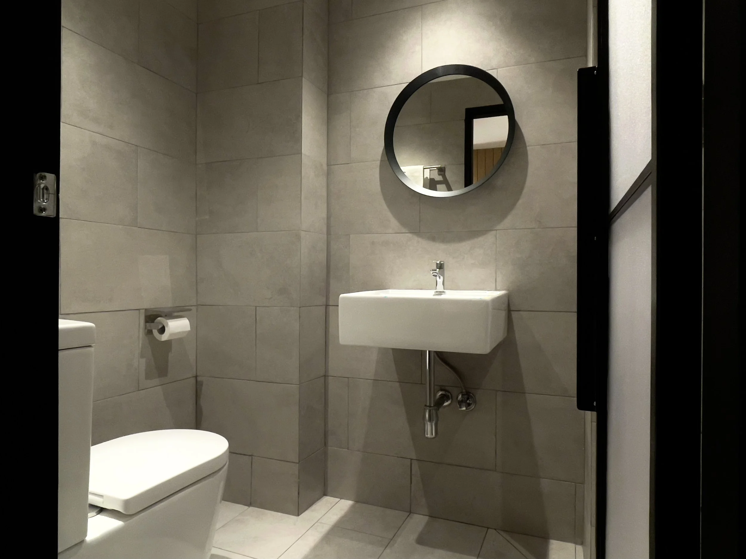 A small bathroom with beige tiled walls, a wall-mounted white sink with a chrome faucet, a round mirror above the sink, a white toilet with a closed lid, and a black door on the right side.