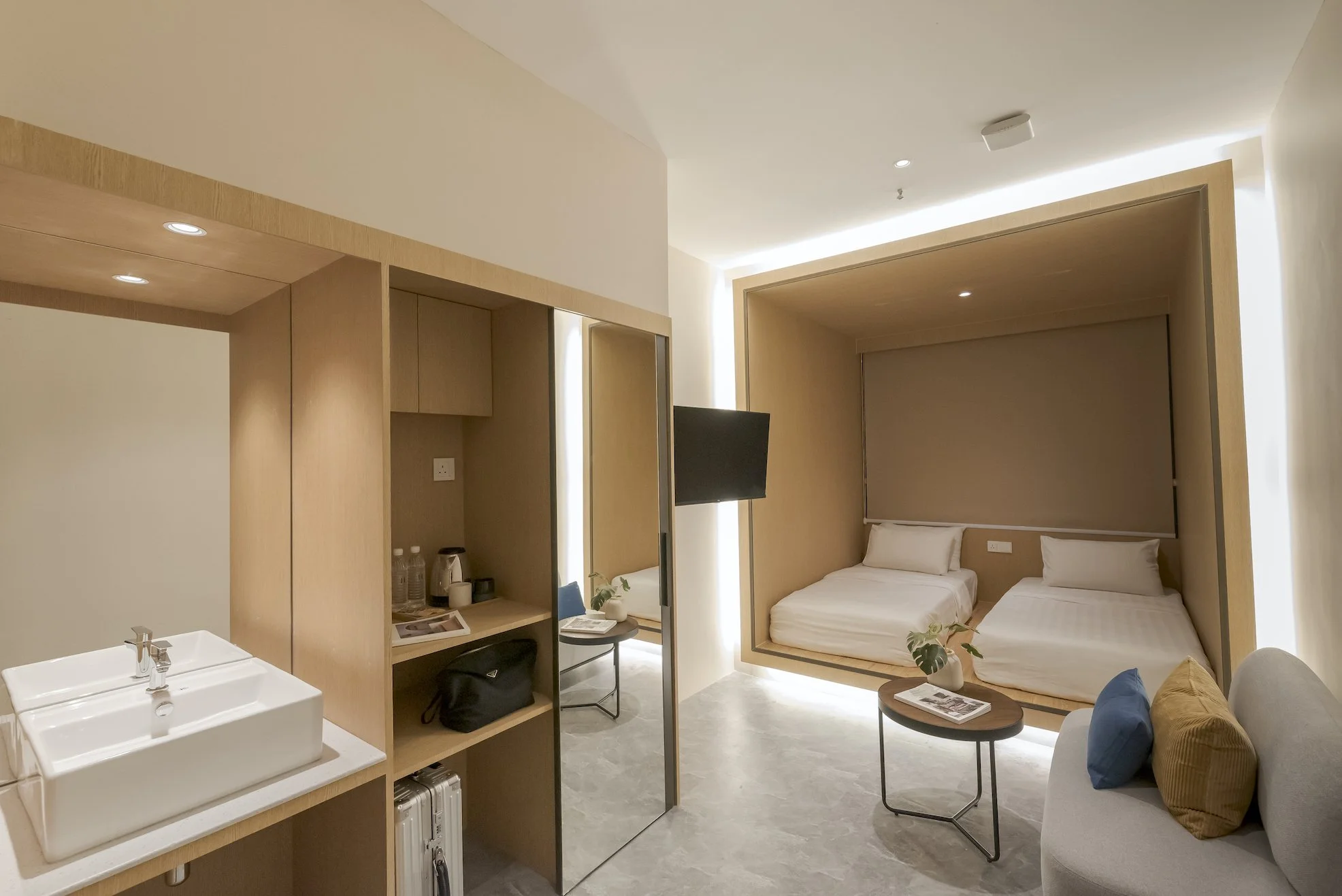 Modern hotel room with two beds in a recessed nook, a small coffee table with a plant and magazine, a sofa with pillows, a wall-mounted TV, and a sink area, all in neutral tones.
