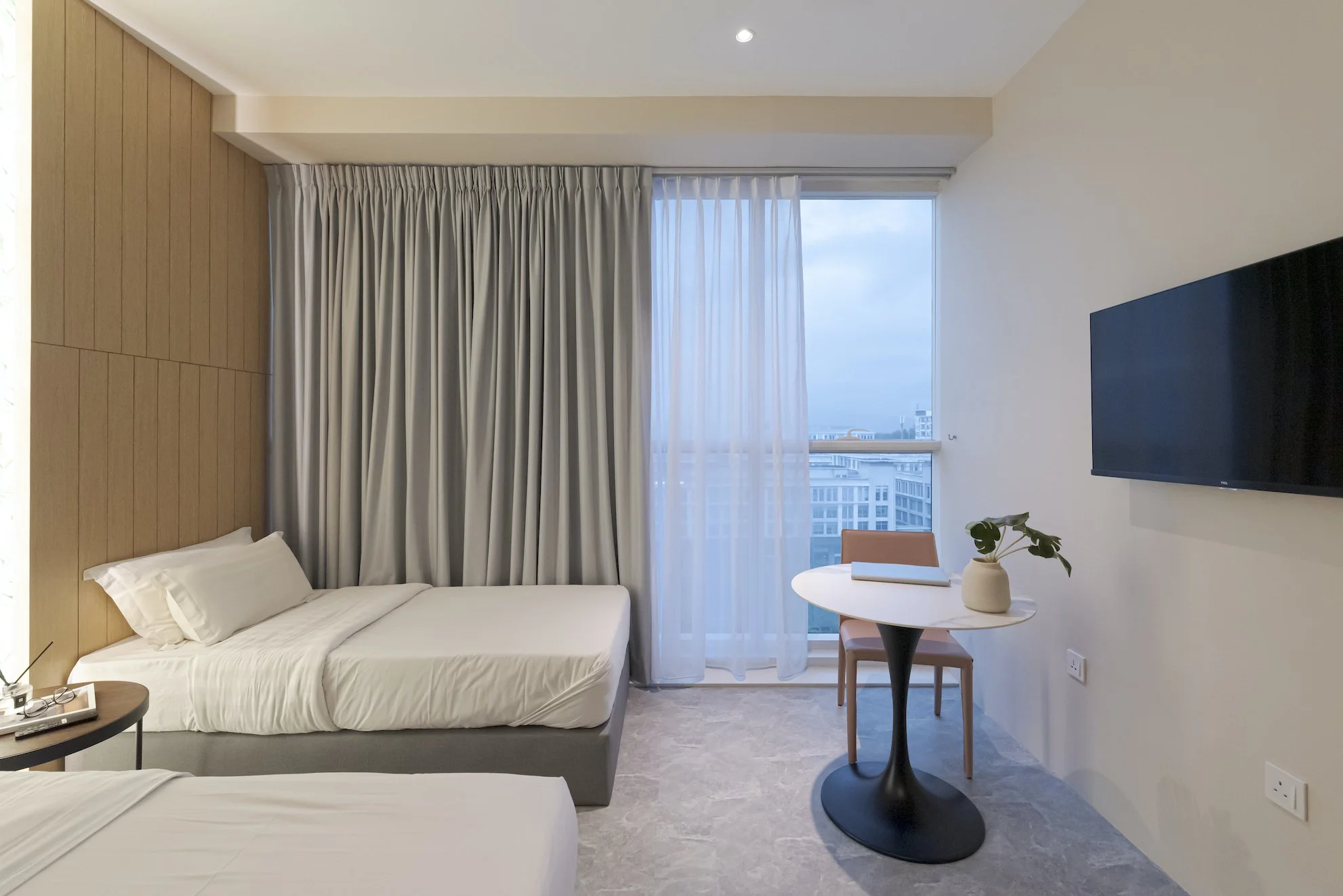 A hotel room with two beds, a small round table with a potted plant and a notebook, a wall-mounted TV, large windows with curtains, and an indoor view of neighboring buildings.