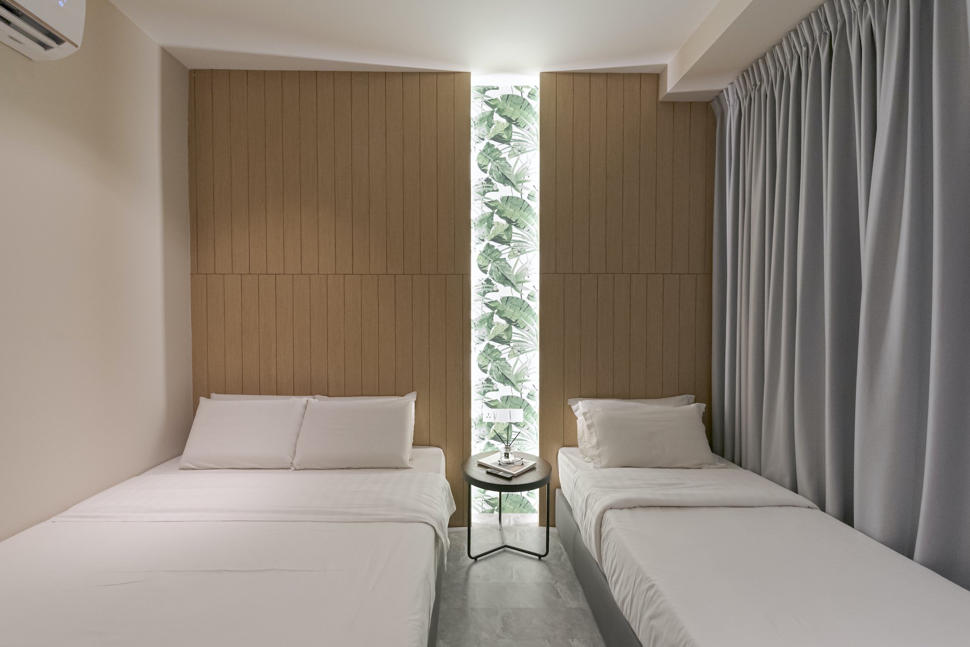 A hotel room with two twin beds, a small round nightstand with a tray, lamp, and book between them, wooden panel wall behind the beds, a vertical window with leafy green plants outside, and light grey curtains on the right.
