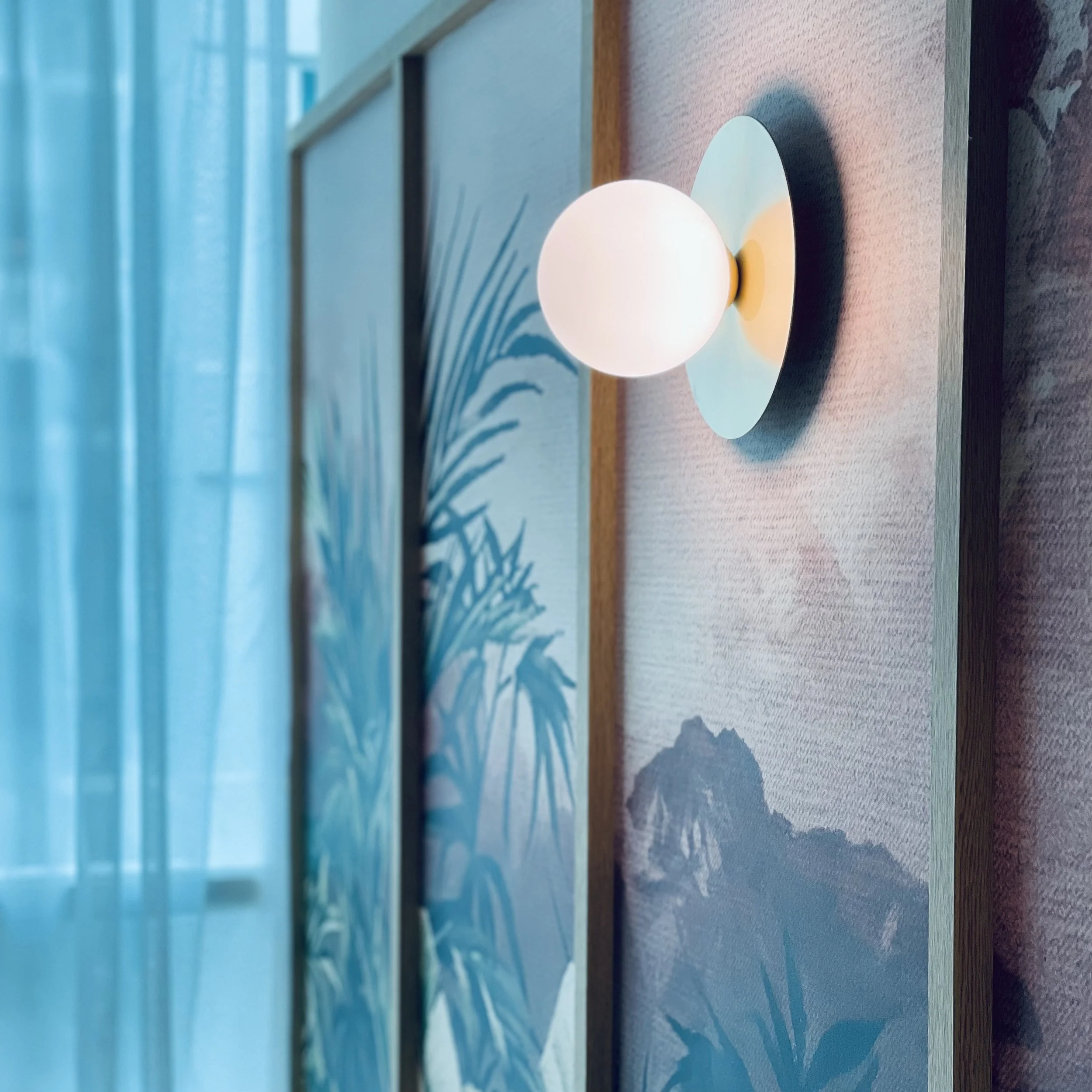 Close-up of a modern wall-mounted lamp with a round white globe shade, attached to a wall with a painted landscape and mountain art in the background.