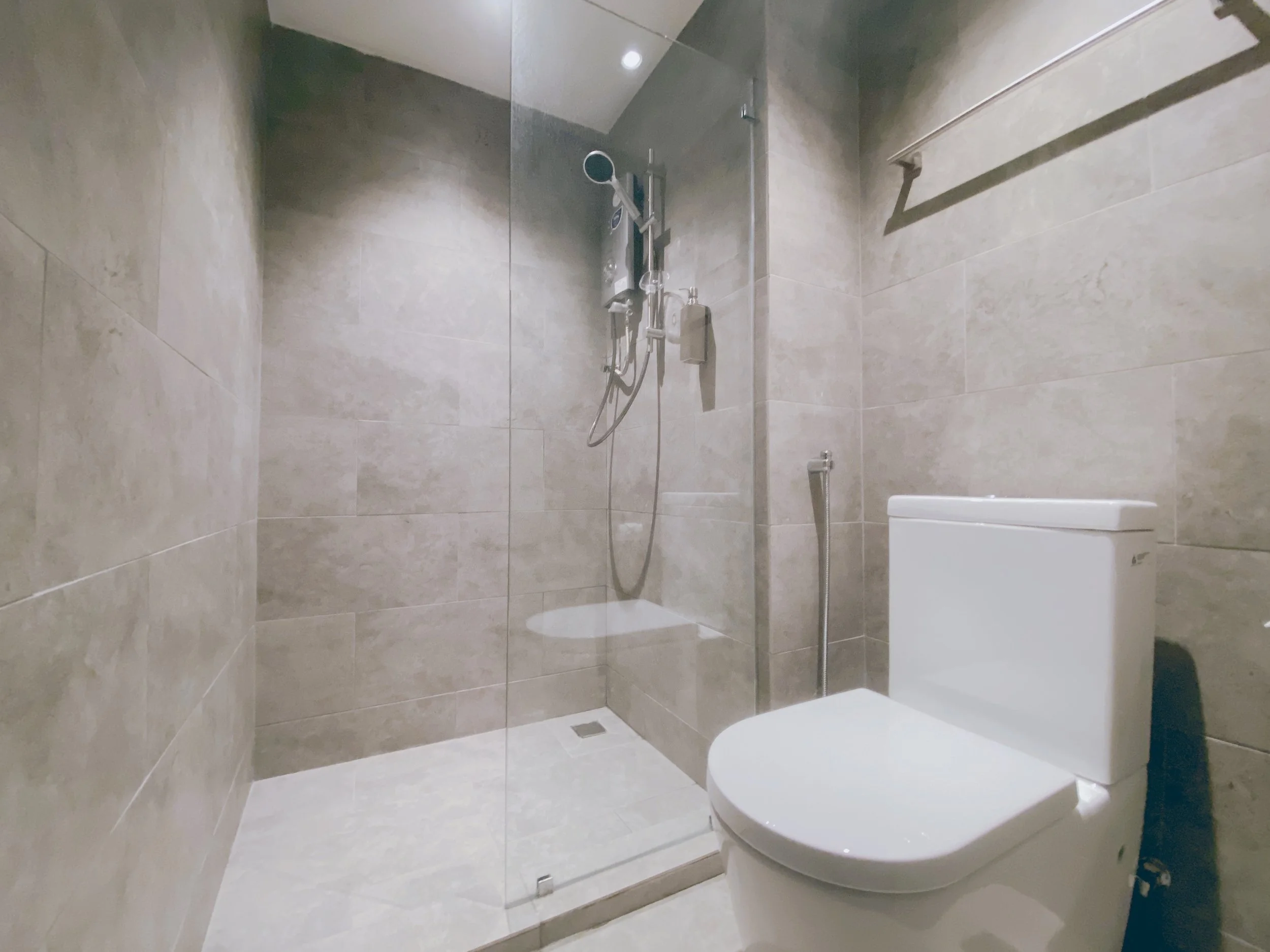 Bathroom with a walk-in shower and a white toilet, beige tiled walls and floor, glass shower door, showerhead and a towel rack on the wall.