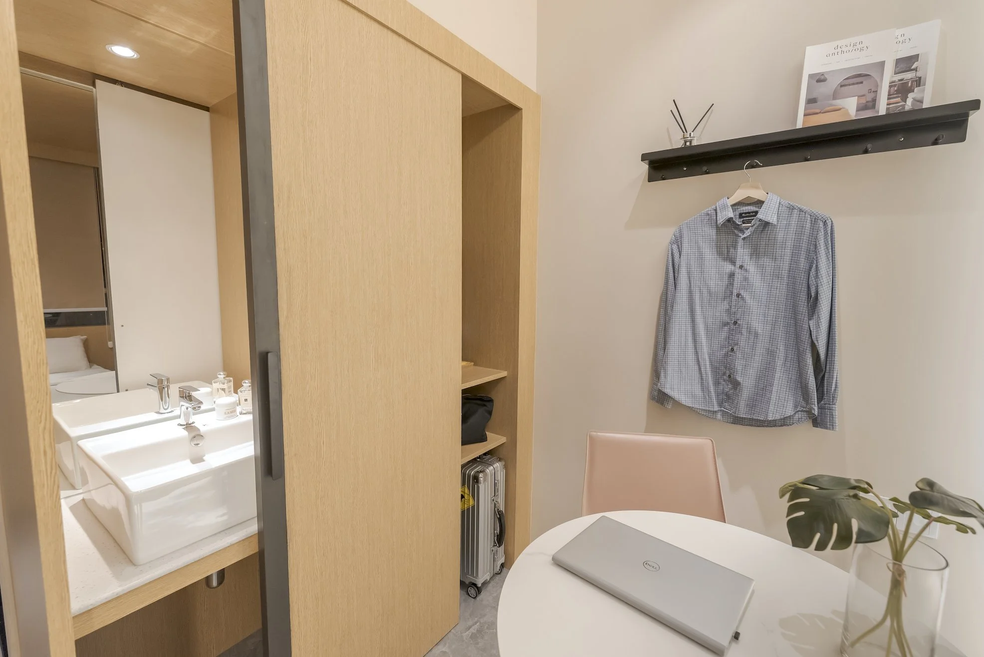 A hotel room with a small round table with a closed silver laptop and a vase with green monstera leaves, a pink chair, a wall shelf with a framed magazine, a gray checkered shirt hanging on a hook, a wooden sliding door, a built-in shelf with a black bag and a briefcase, and a sink area with two white square sinks, soap dispensers, and a mirror.