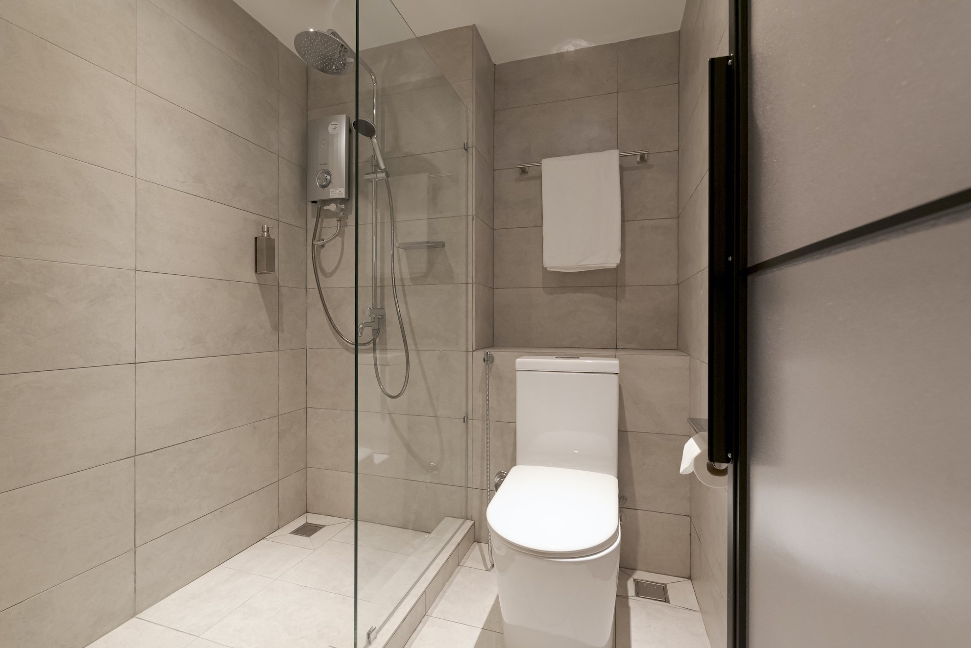 Modern bathroom with walk-in shower, rainfall showerhead, white toilet, beige tiled walls and floor, white towel hanging on a rail.